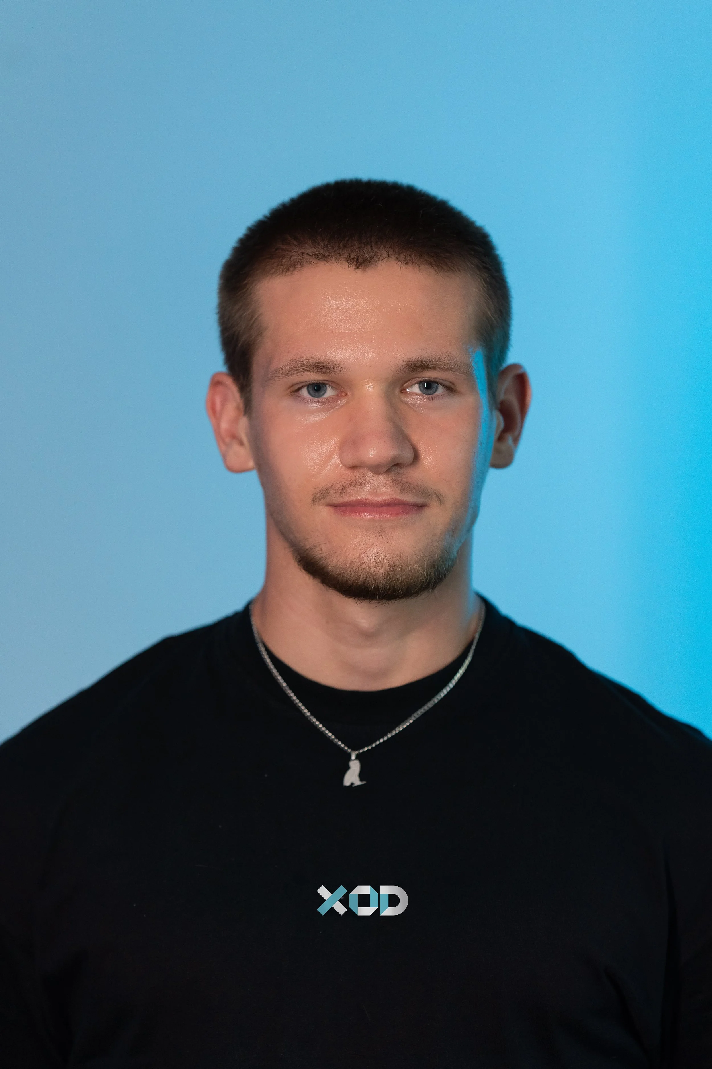 Young man with short hair and a goatee, wearing a black shirt with the letters 'XOD' on it and a silver necklace with a penguin pendant, standing against a blue background.