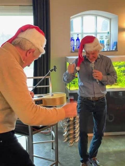 Two men wearing Santa hats are dancing and having fun, with one playing a guitar. They are indoors near a window with a view of a garden.