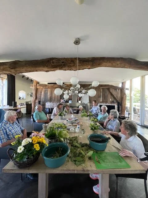 Elderly people sitting around a dining table decorated with flowers in a bright room with large windows and a wooden ceiling beam.