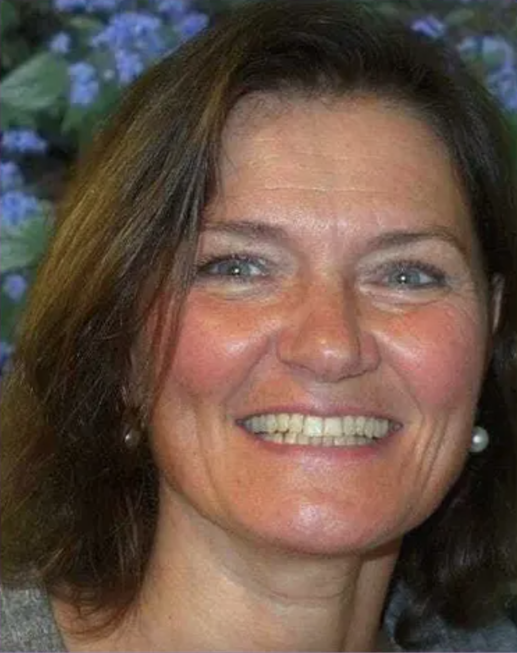 A woman with blue eyes, brown hair, and a bright smile showing her teeth, wearing pearl earrings, with a background of blue flowers.