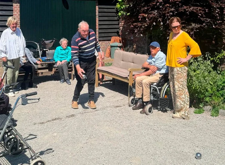 Group of elderly people and caregivers playing bocce ball outdoors in a garden patio area.