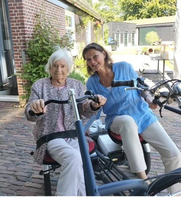 An elderly woman and a middle-aged woman sitting on pedal bikes outdoors, smiling at the camera. The background includes a brick house and a garden.