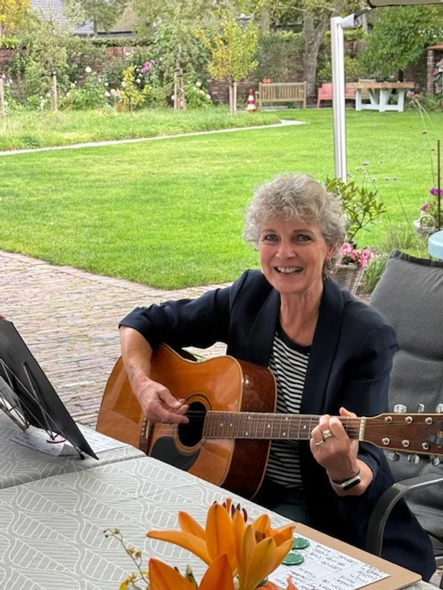 A woman sitting outdoors playing an acoustic guitar and smiling, with a garden and patio in the background.