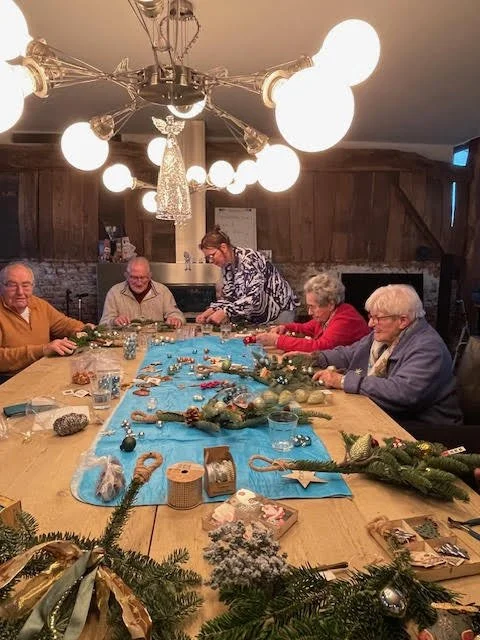 A group of elderly people sitting around a table decorated for Christmas, with a woman arranging ornaments. The table has a blue table runner, Christmas decorations, and holiday-themed items. A modern chandelier with multiple globe lights hangs above.