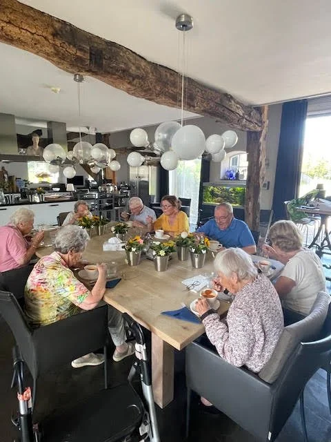 Elderly people gathered around a dining table, eating and chatting in a modern, well-lit room with hanging globe lights and large windows.