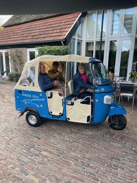 Three people sitting inside a blue and cream tuk-tuk parked in front of a building with large glass windows.