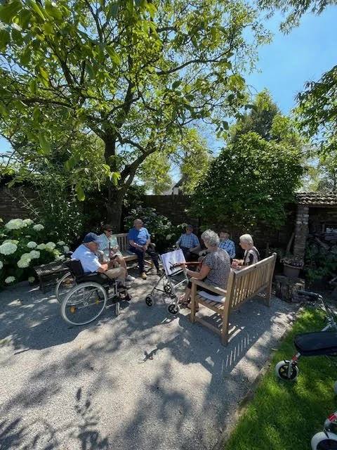 Group of elderly people sitting outdoors around a table and on benches under a large tree in a garden.
