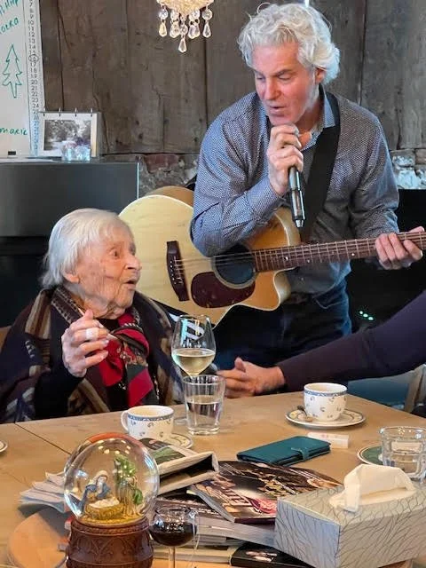 An elderly woman sitting at a table with a glass of white wine, engaging with a man playing a guitar and singing. There are cups, glasses, magazines, and a snow globe on the table. The setting appears cozy and warm.