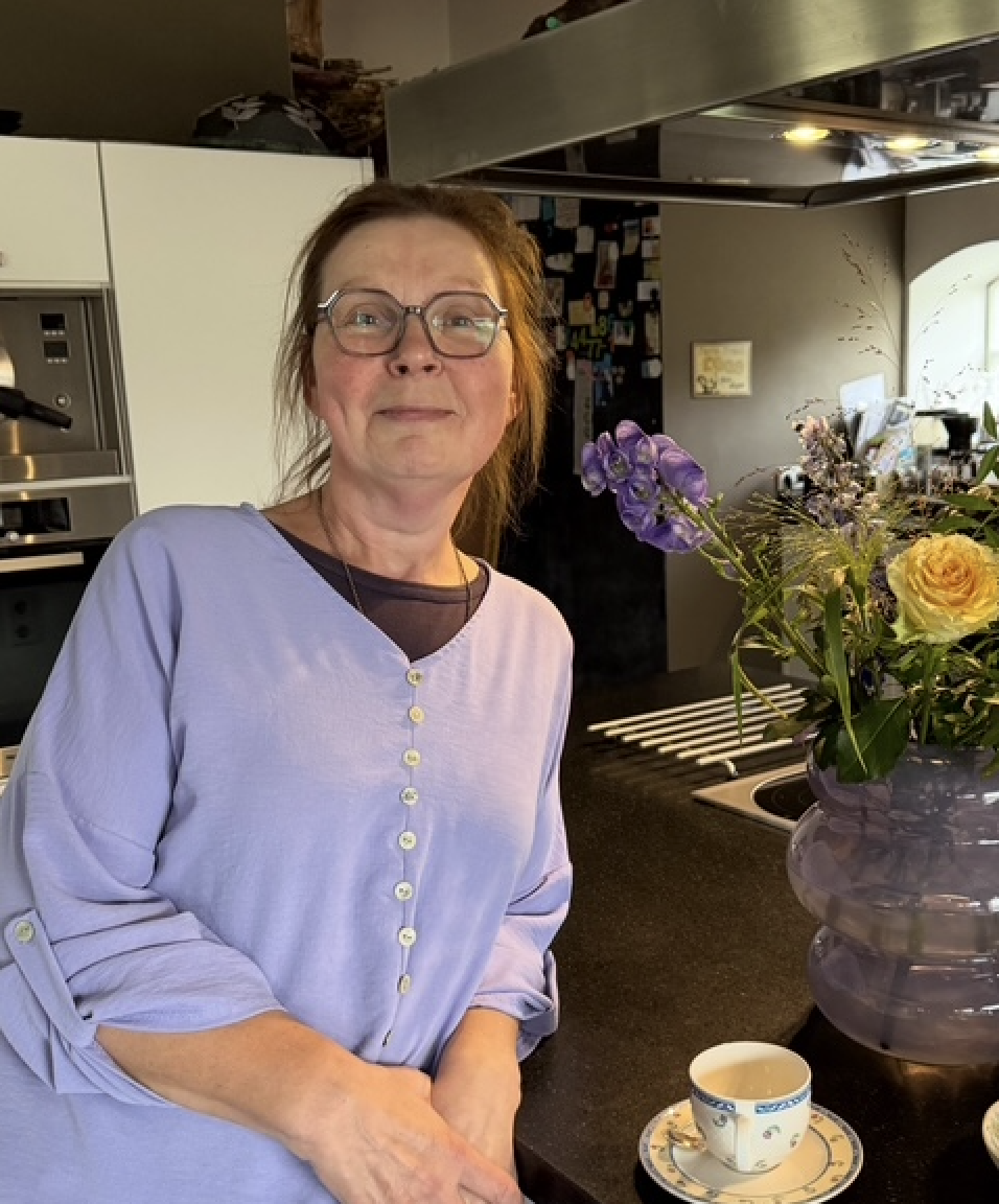 A woman with glasses and brown hair stands in a kitchen, smiling, with a large purple vase of flowers and a cup on the counter.