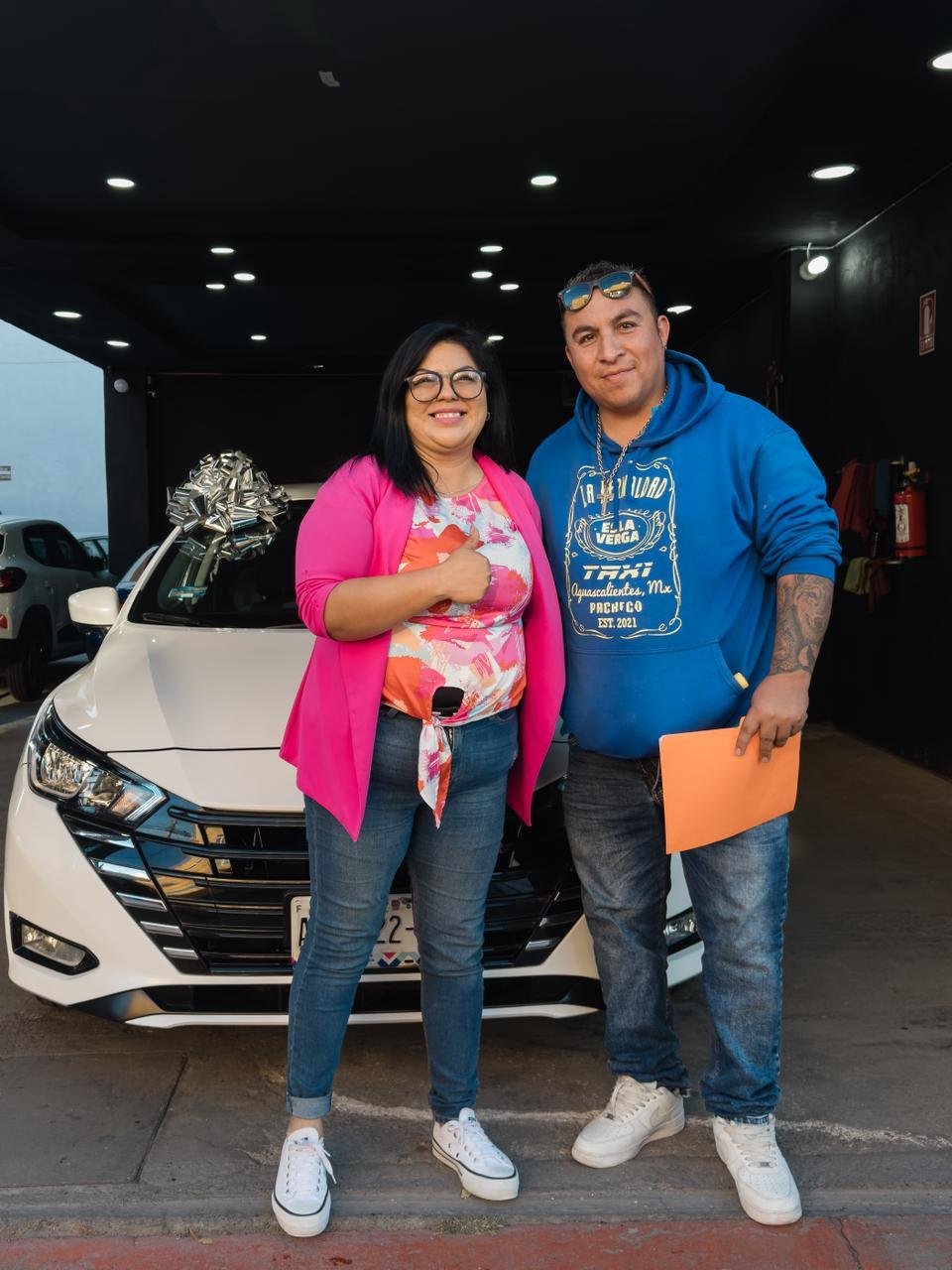UNA MUJER Y UN HOMBRE SONRIENTES DE PIE DELANTE DE UN COCHE BLANCO, CON UN GRAN LAZO PLATEADO SOBRE EL COLCHÓN DEL VEHÍCULO, EN UN ÁREA CUBIERTA CON LUCES SOBRE EL TECHO.