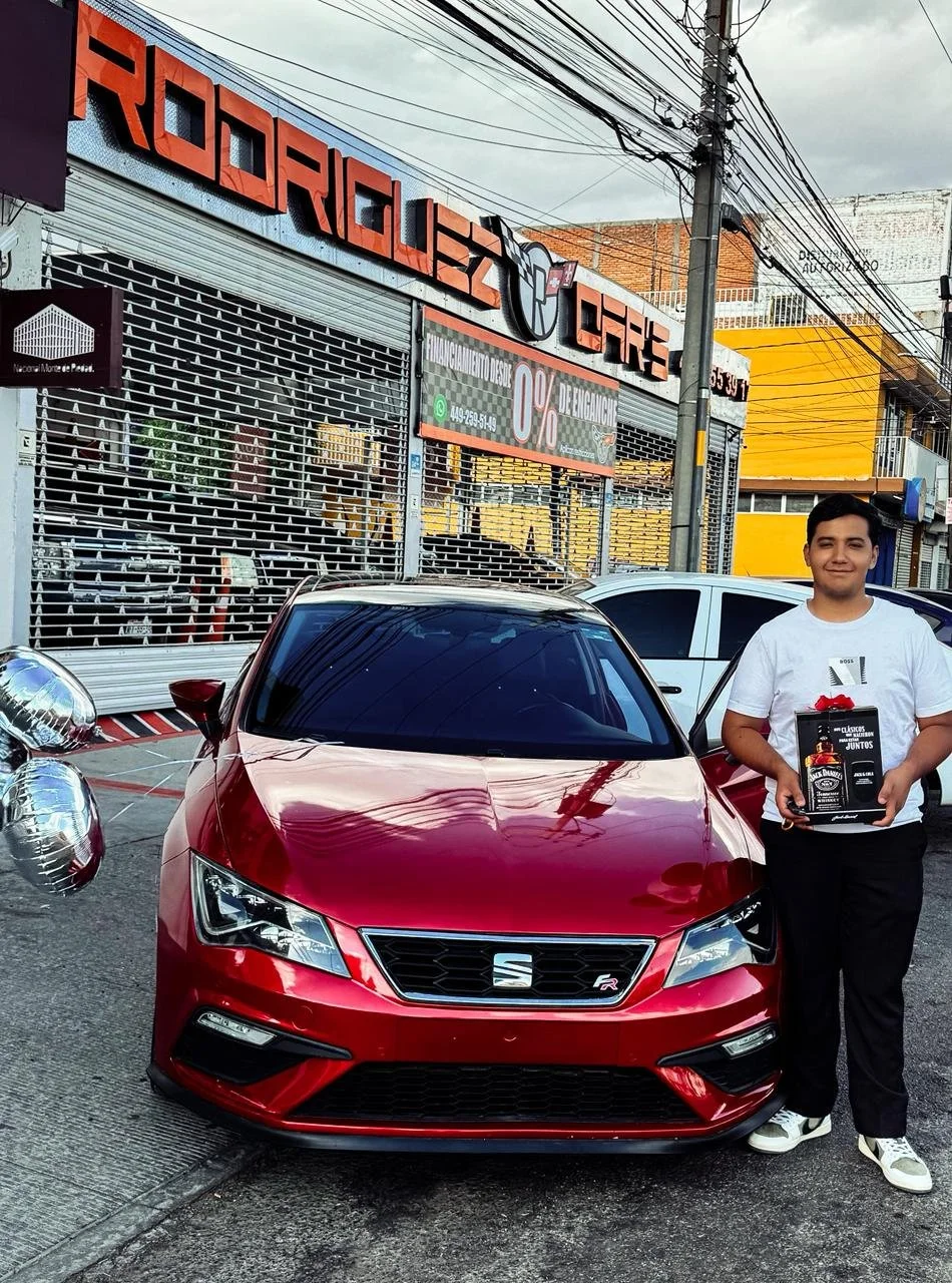 Joven sonriendo sosteniendo un paquete con el logotipo de la marca de autos SEAT, de color rojo, parada frente a un auto de la misma marca y modelo en la calle, con locales comerciales y postes de electricidad en el fondo.