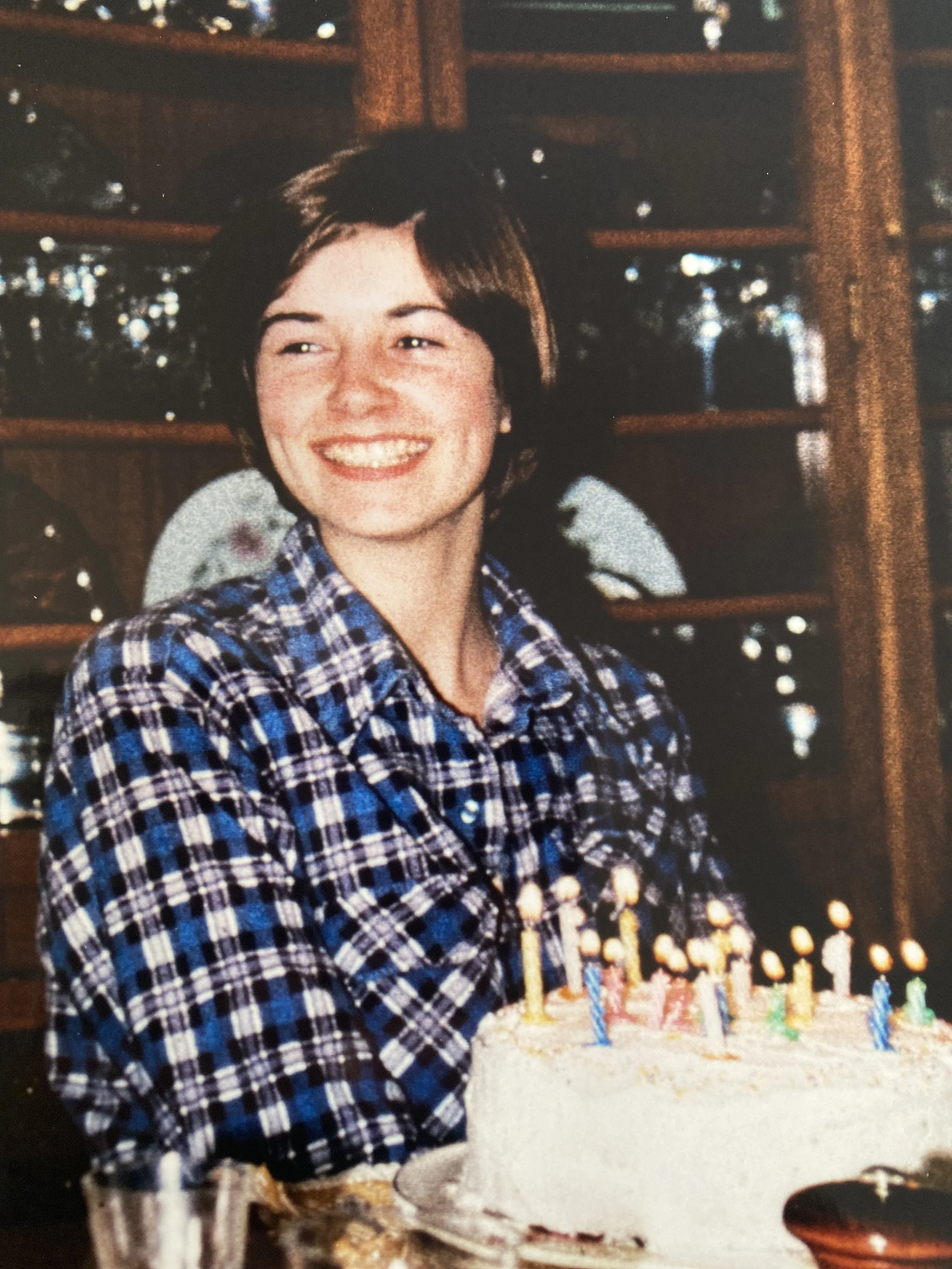 A young woman with short brown hair, smiling, wearing a blue plaid shirt, sitting behind a birthday cake with lit candles.