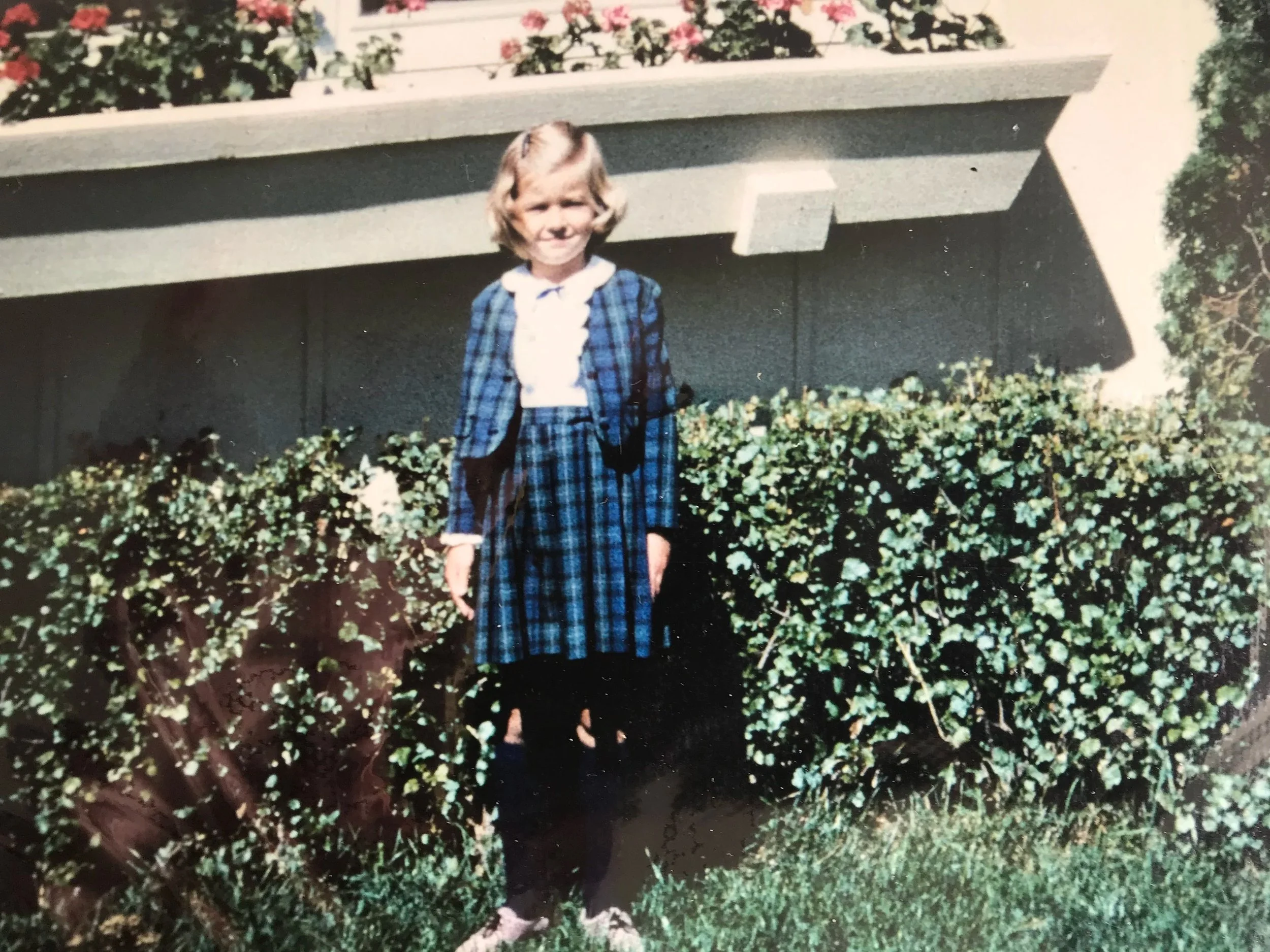 A young girl stands in front of a bush with a house behind her. She is wearing a blue plaid school uniform and smiling at the camera.