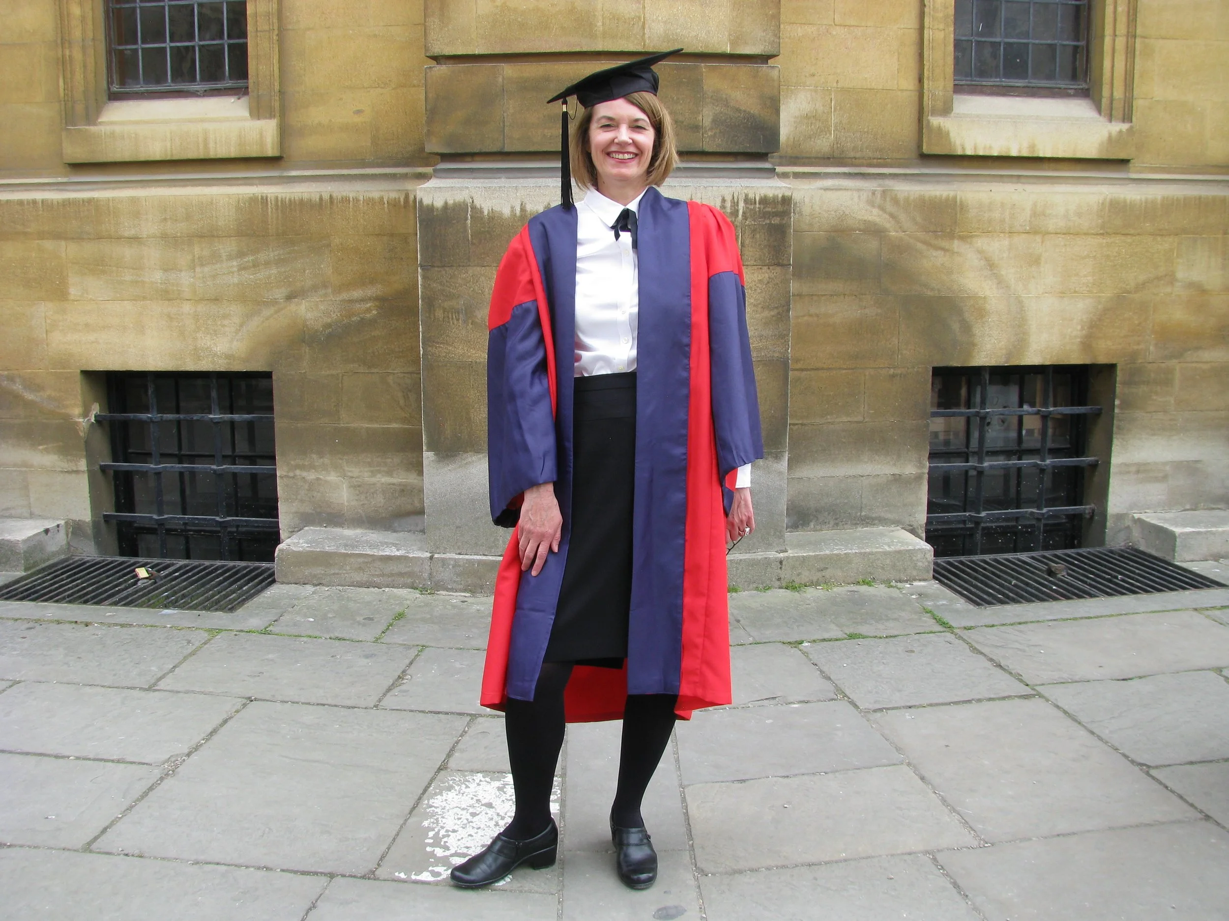 A smiling woman in graduation attire stands outside a building with stone walls and barred windows.
