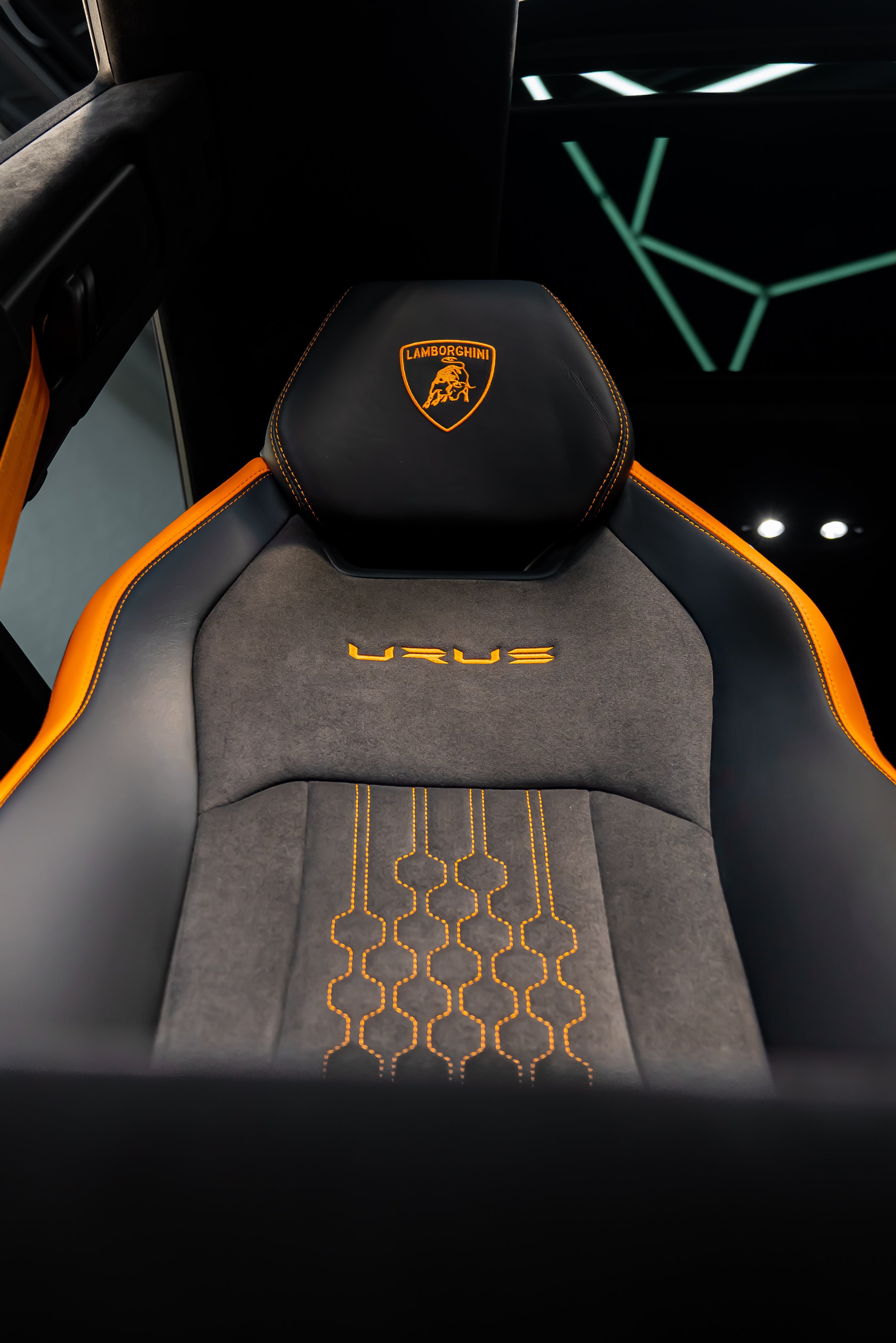 The interior of a Lamborghini Urus, featuring a black leather seat with orange stitching, Lamborghini logo on the headrest, and orange embroidered text 'URUS' on the seat cushion. The background shows a modern, illuminated ceiling with a geometric pa