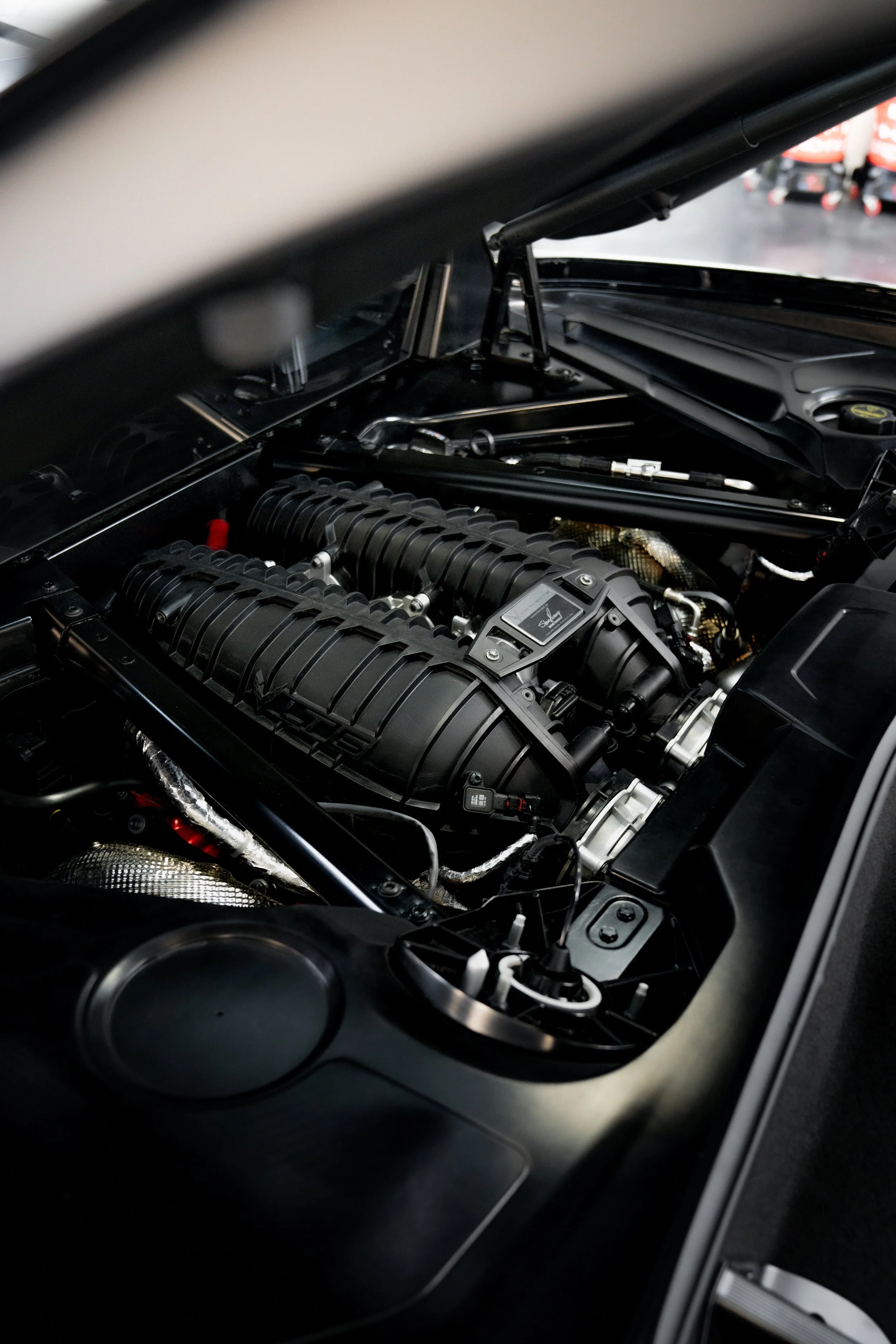 Close-up of the car engine inside the hood of a C8 Chevrolet Corvette Z06 with visible black intake manifolds and various engine components.