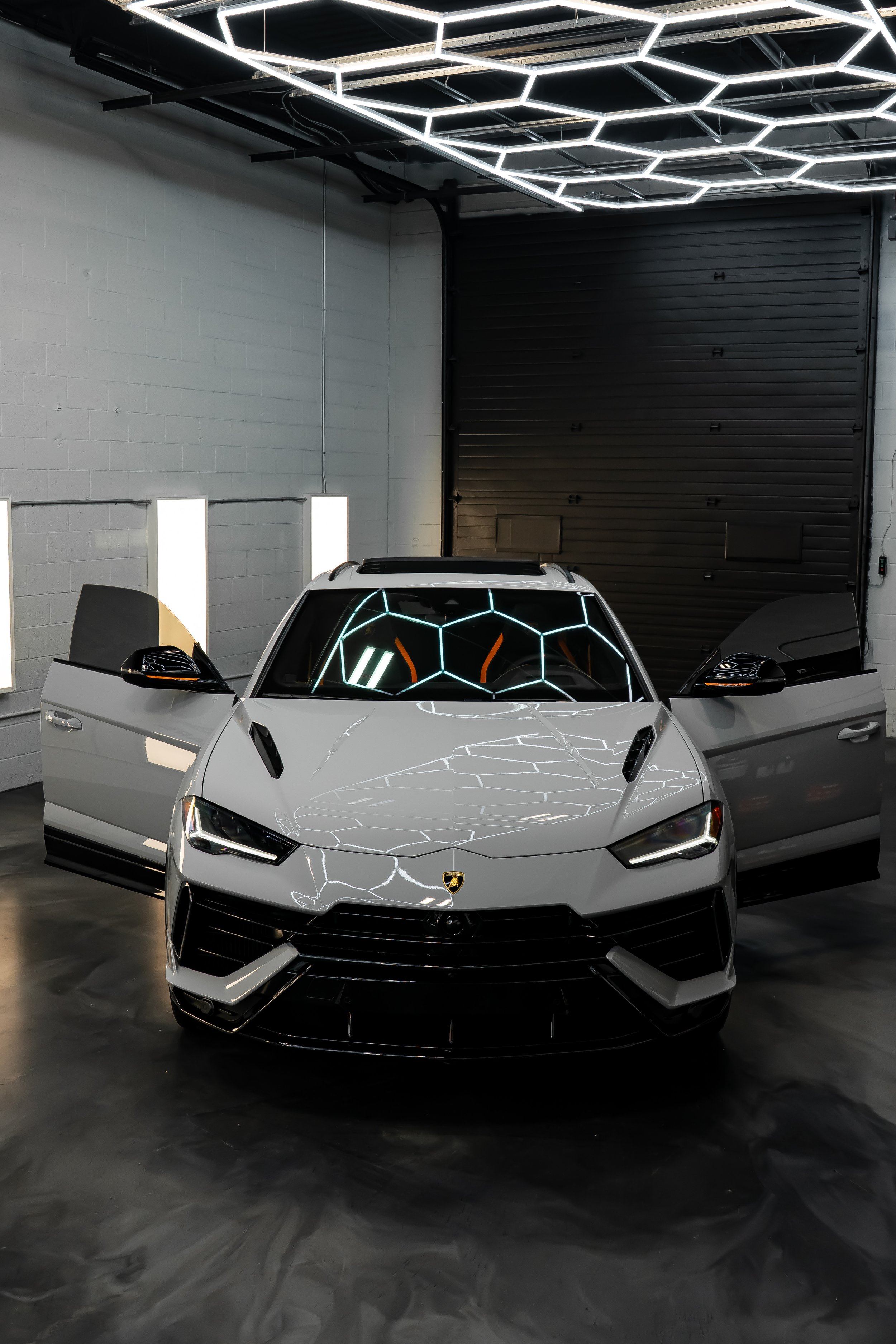 A grey Lamborghini Urus with open doors parked in ZAutoCare's wash bay with a hexagon light ceiling and a black garage door in the background.