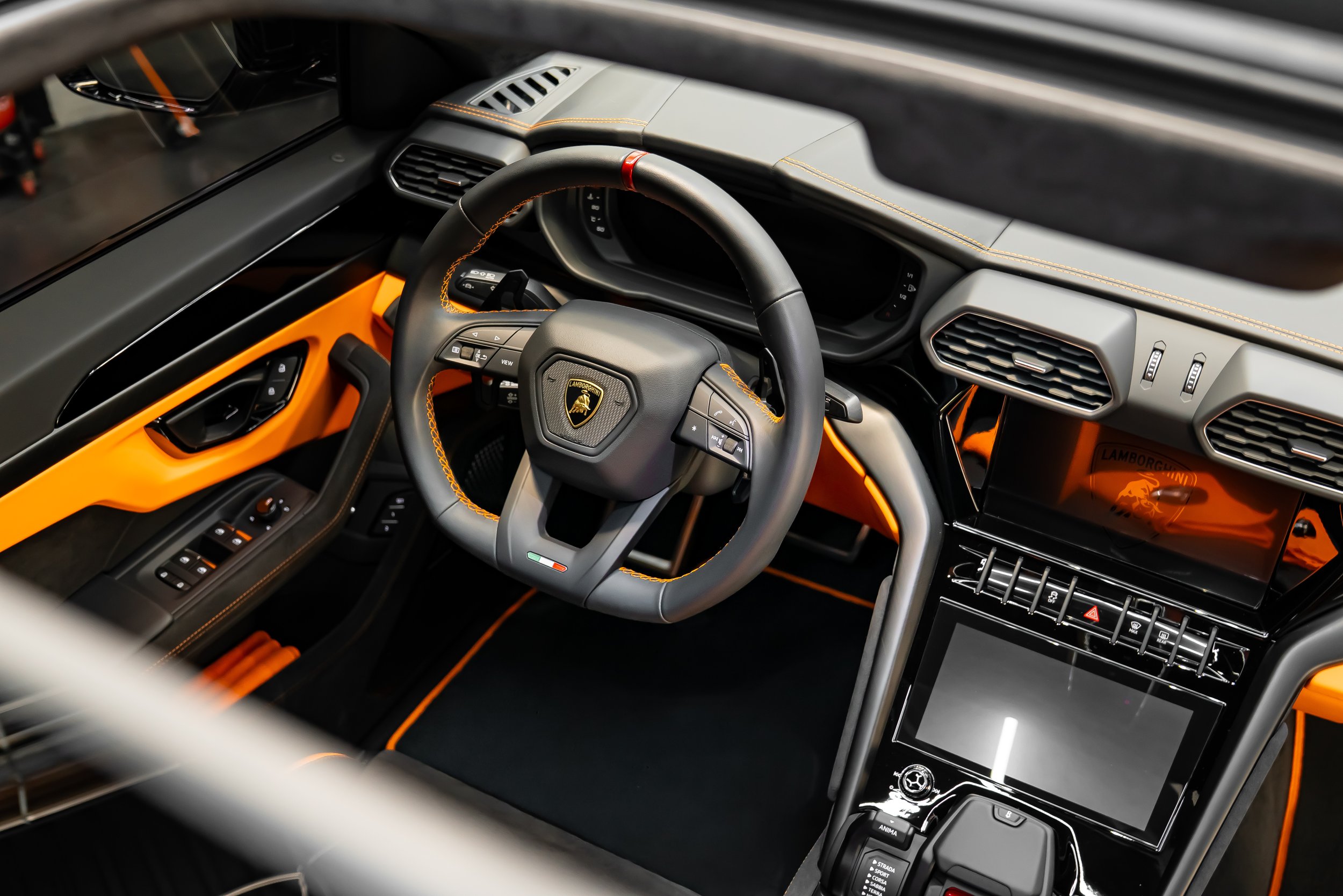 Interior of a Lamborghini supercar, featuring a sporty steering wheel with the Lamborghini emblem, orange accents, and modern dashboard with digital displays.
