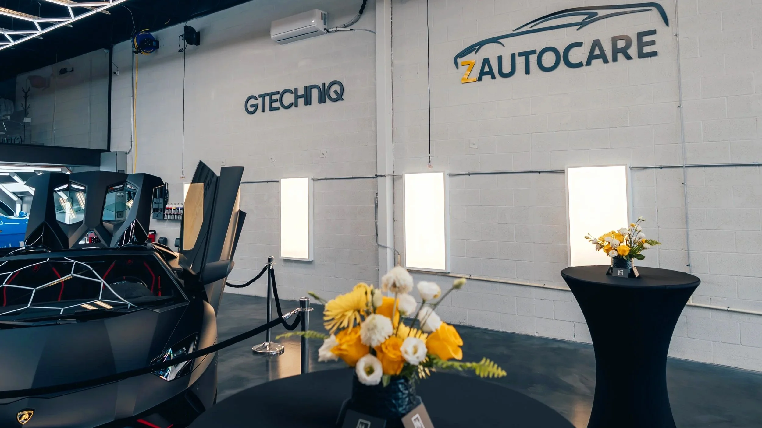 An indoor look of ZAutoCare's facility featuring the ZAutoCare and Gtechniq logo on the wall with a black Lamborghini Aventador Ultimae Roadster and Porsche GT3 RS on display.