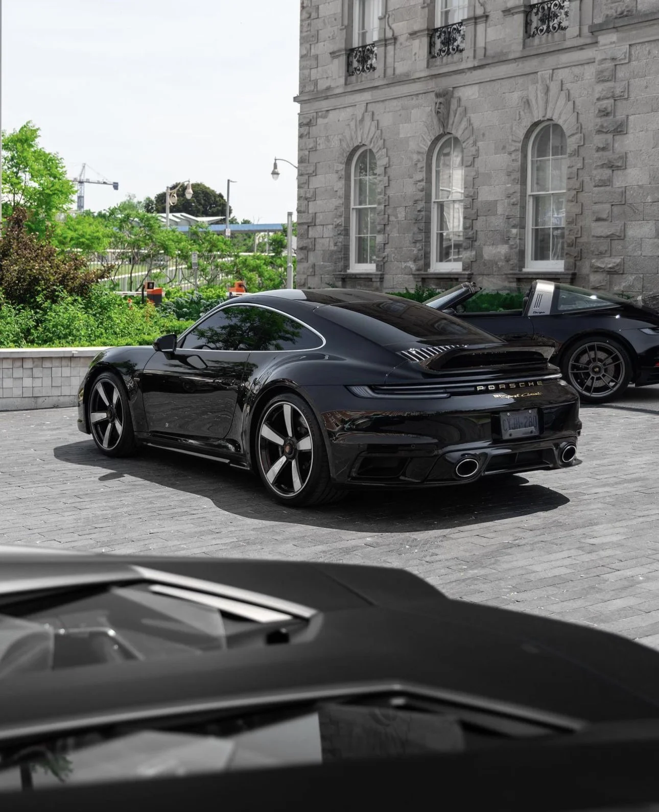 A rare Black Porsche 911 Sports Classic sports car parked on a cobblestone street with historic stone building and another sports car in the background in Guelph.