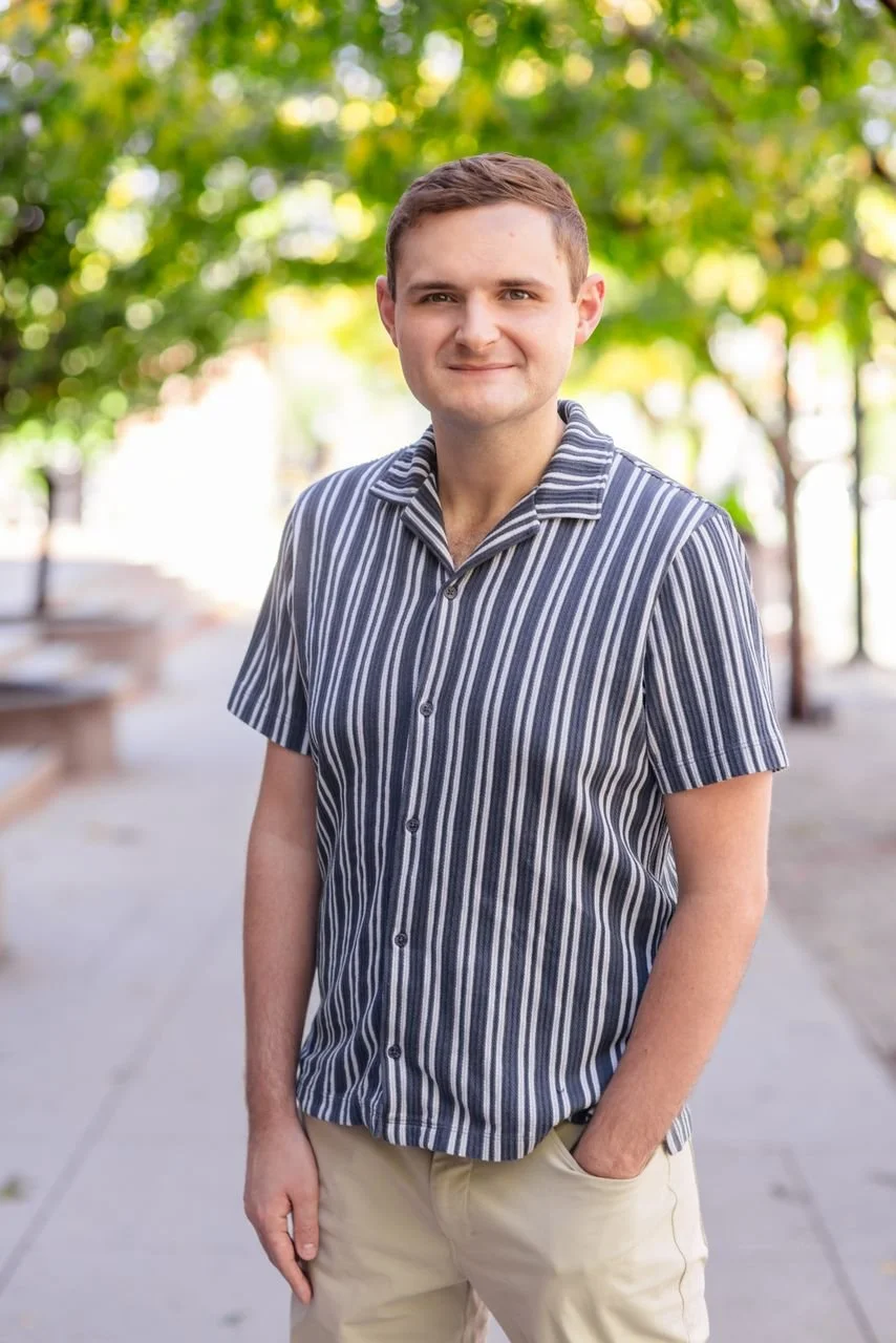 A young man with short brown hair and fair skin, wearing a short-sleeved, vertically striped shirt in shades of blue, gray, and white, stands outdoors with a background of green trees and blurred park benches.