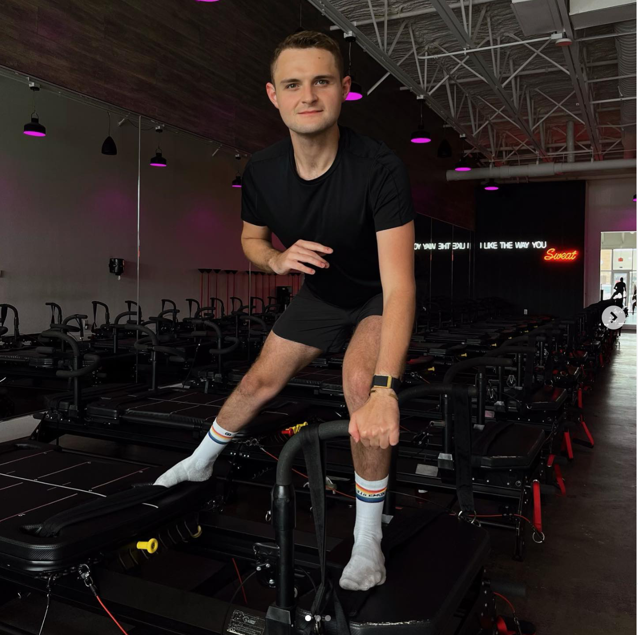 A man in black athletic wear and white socks on a mini treadmill at a fitness studio, with purple lighting and a neon sign in the background.