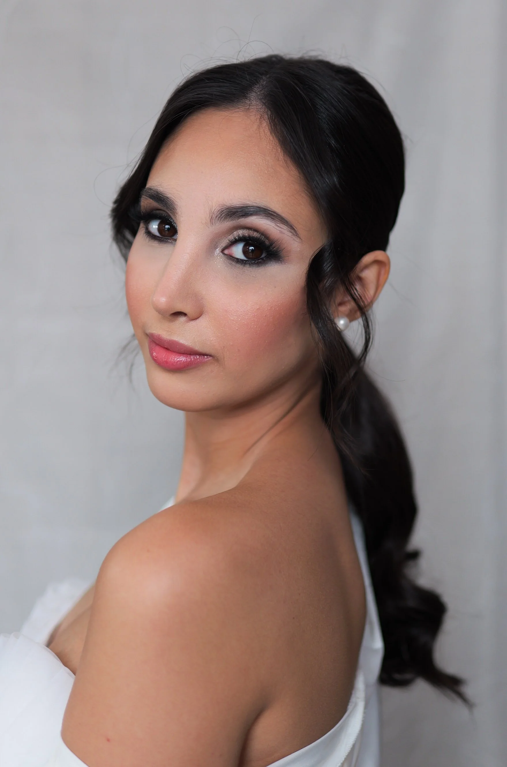 Close-up portrait of a young woman on her wedding day with dark hair, makeup, and pearl earrings, looking at the camera against a neutral background.