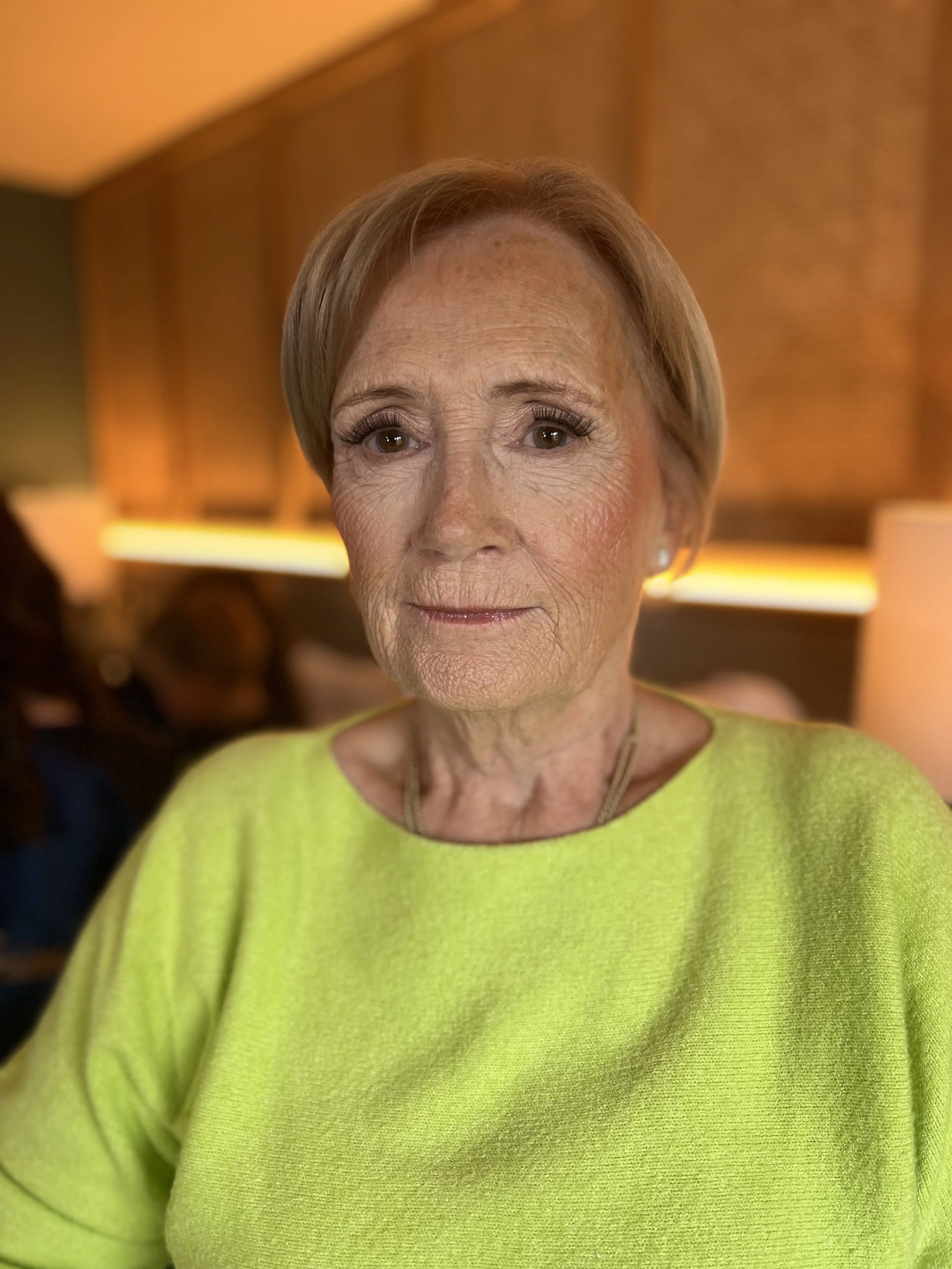 An elderly woman with short gray hair smiling softly, wearing a bright yellow-green sweater, in a warmly lit indoor setting with wooden paneling in the background.
