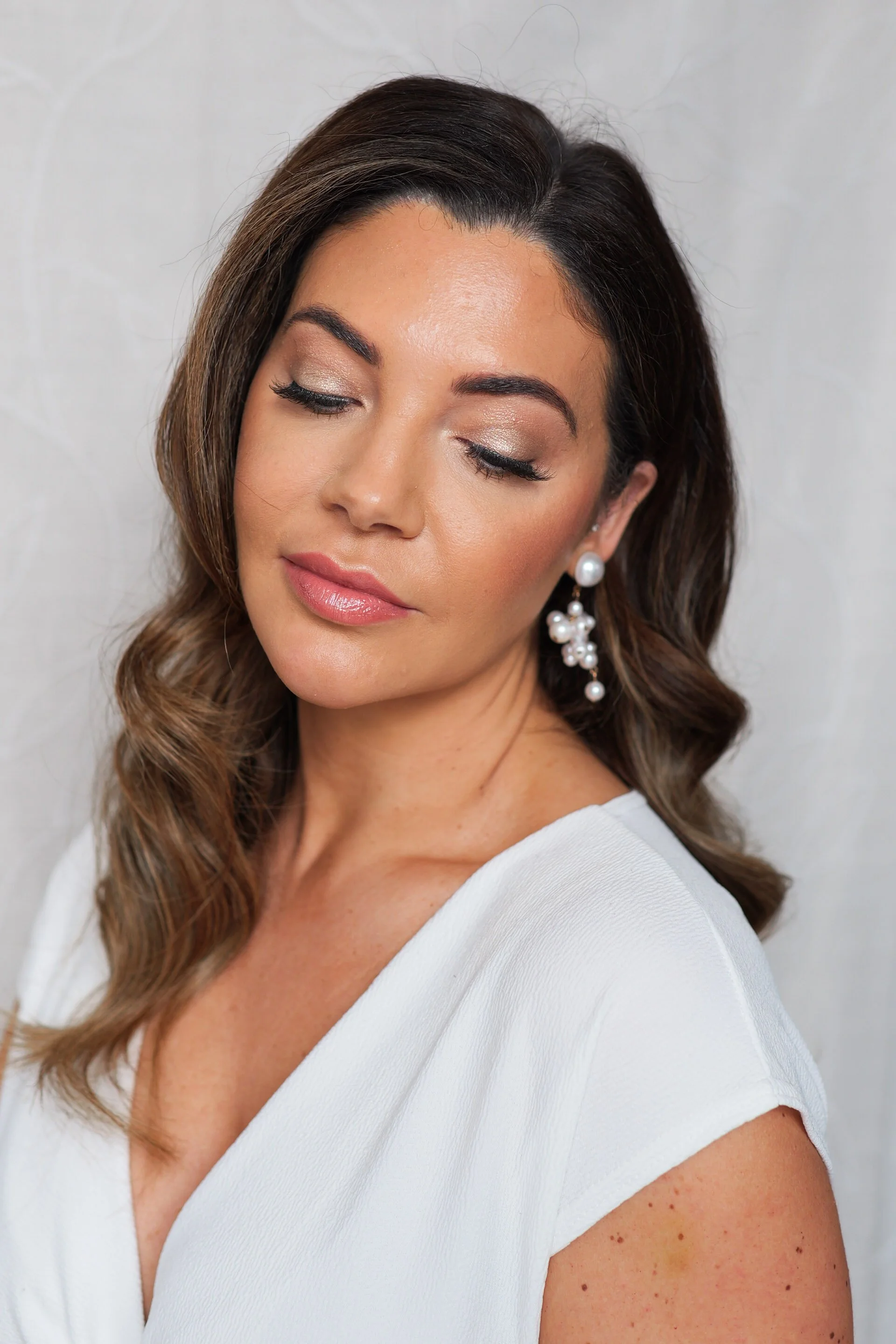 A woman with long, wavy brown hair styled to one side, wearing pearl drop earrings and a white top, with bridal eye makeup and lipstick, against a plain background on her wedding day.