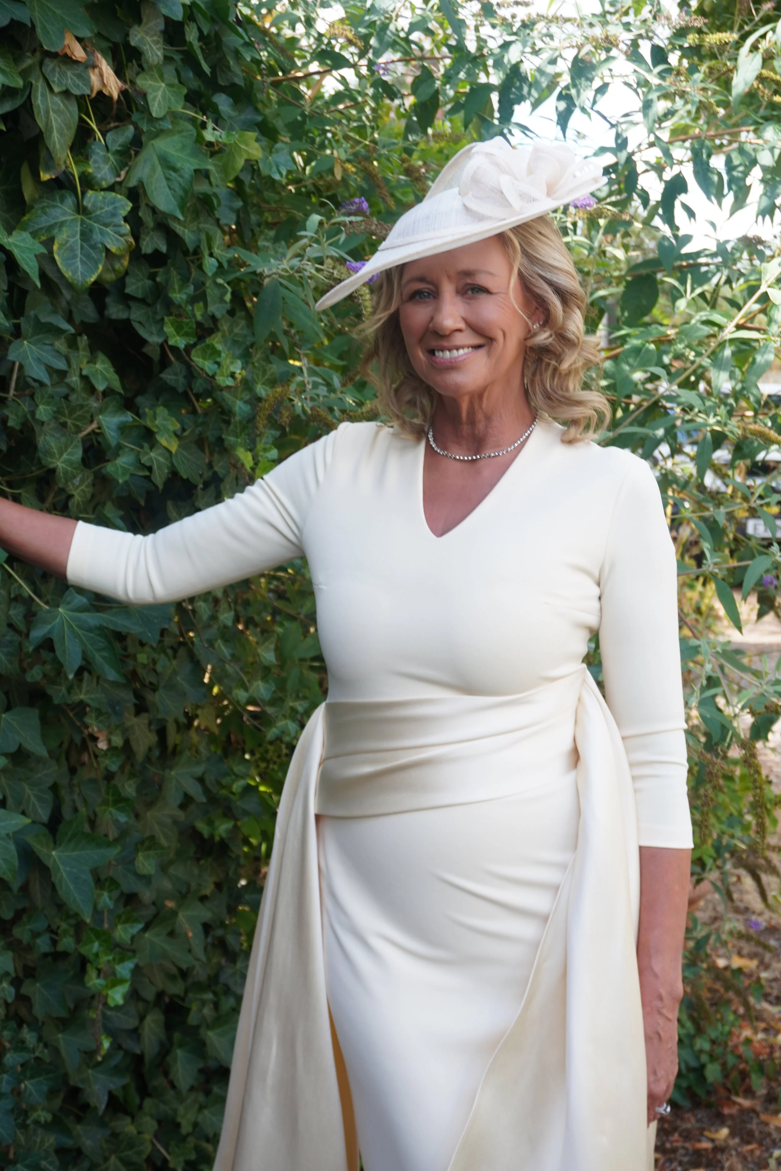 A woman dressed in a cream-colored gown and a matching wide-brimmed hat stands outdoors surrounded by green ivy and plants, smiling at the camera.