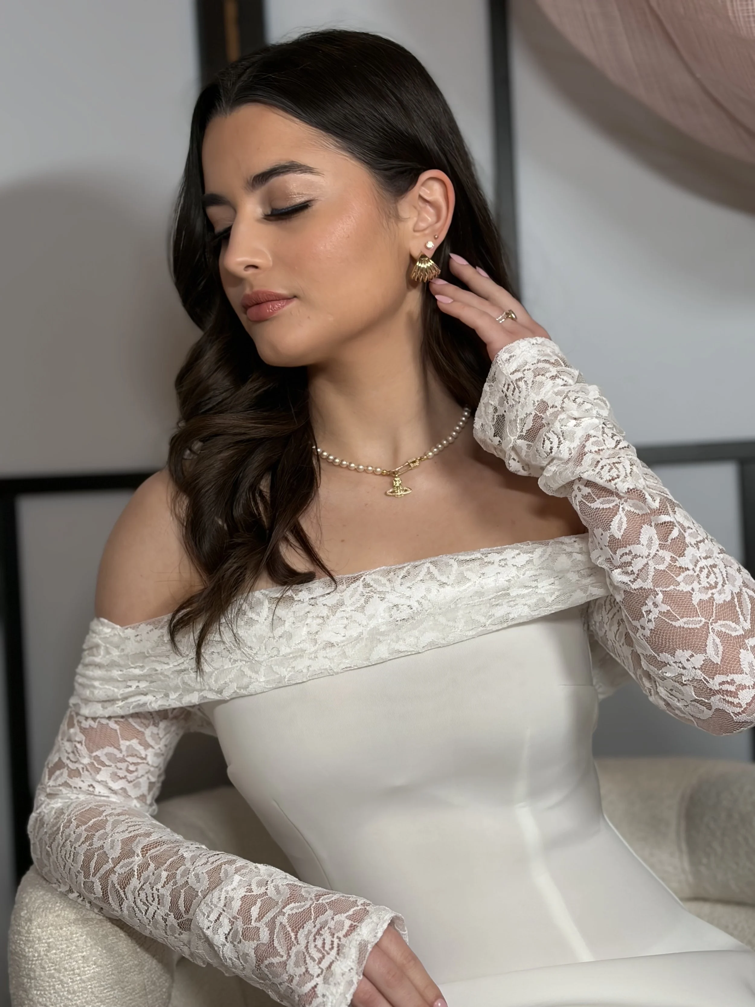A woman in a white off-shoulder lace wedding dress with a pearl necklace and gold jewelry, posing with her eyes closed and her hand touching her neck.