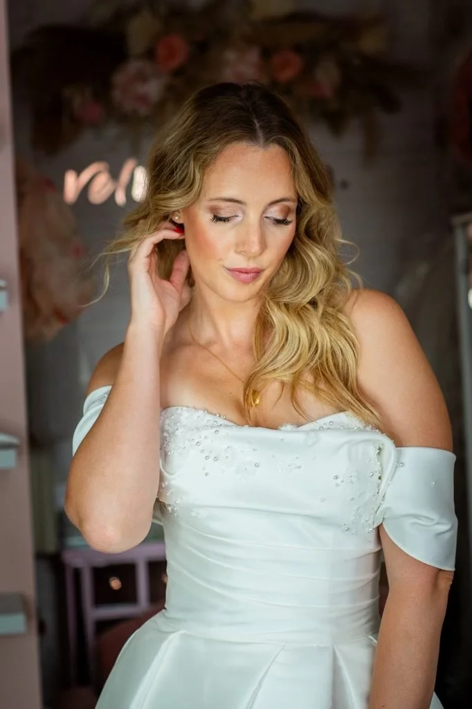 A woman in a white wedding dress with off-the-shoulder sleeves, standing indoors with a floral background, looking downward with her eyes closed and touching her hair.