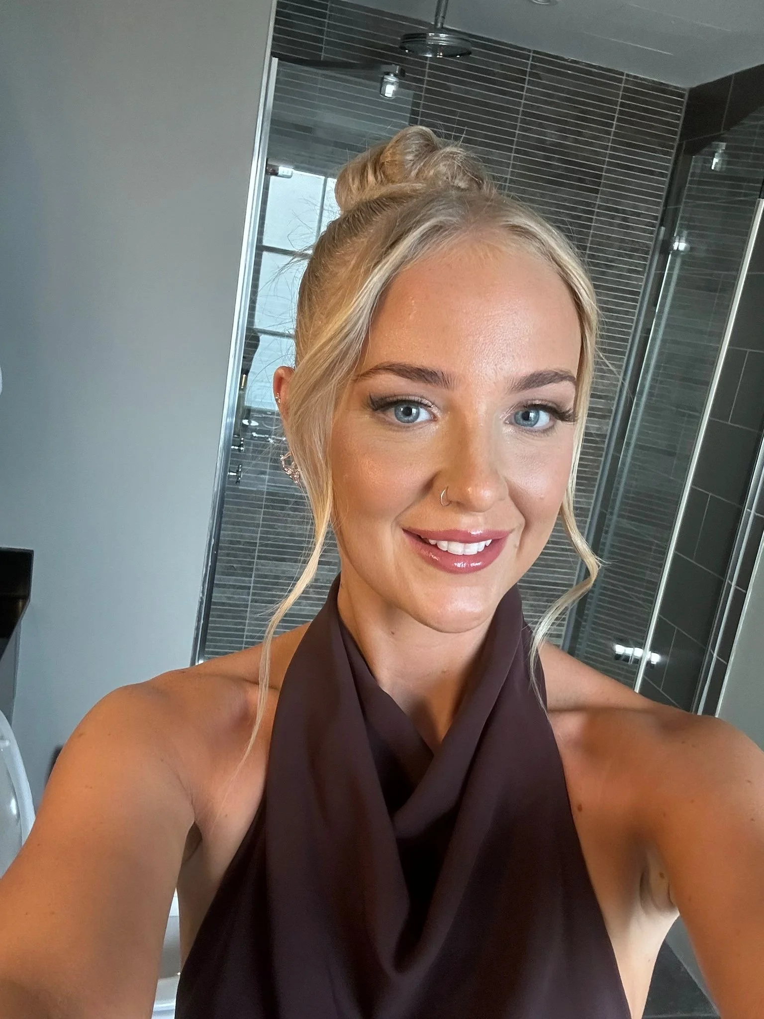 A young woman with blonde hair styled in an updo, wearing a sleeveless dark brown top, taking a selfie in a modern bathroom with black tiled walls and a glass shower.