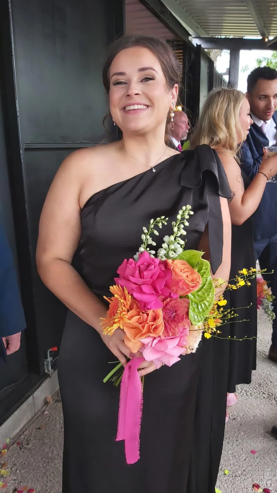 A woman in a black dress holding a colorful bouquet of flowers, smiling at a celebration or event, with other guests in the background.