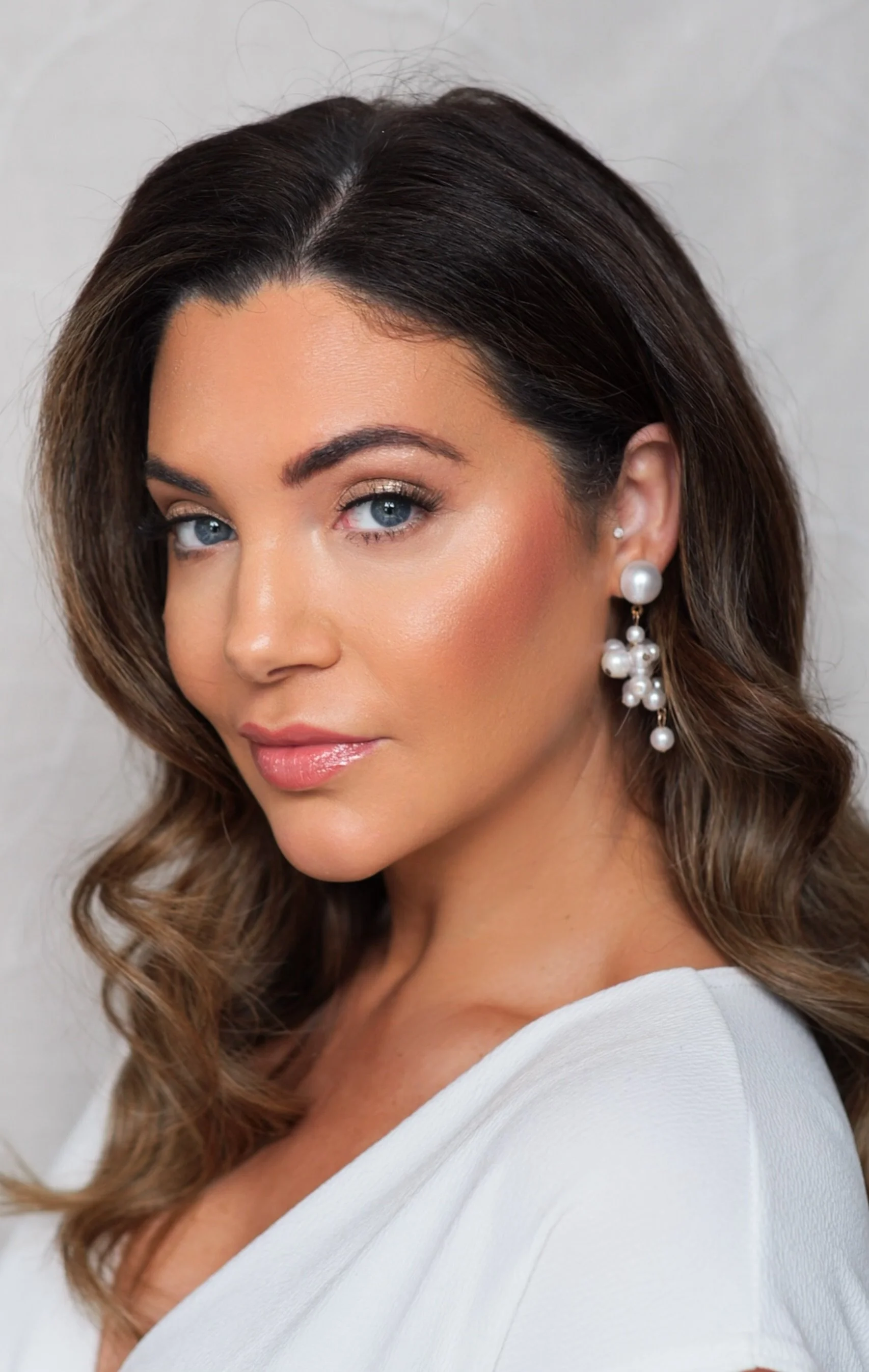 A woman with wavy brown hair, blue eyes, and light makeup, wearing pearl earrings and a white top, looking slightly to the side.