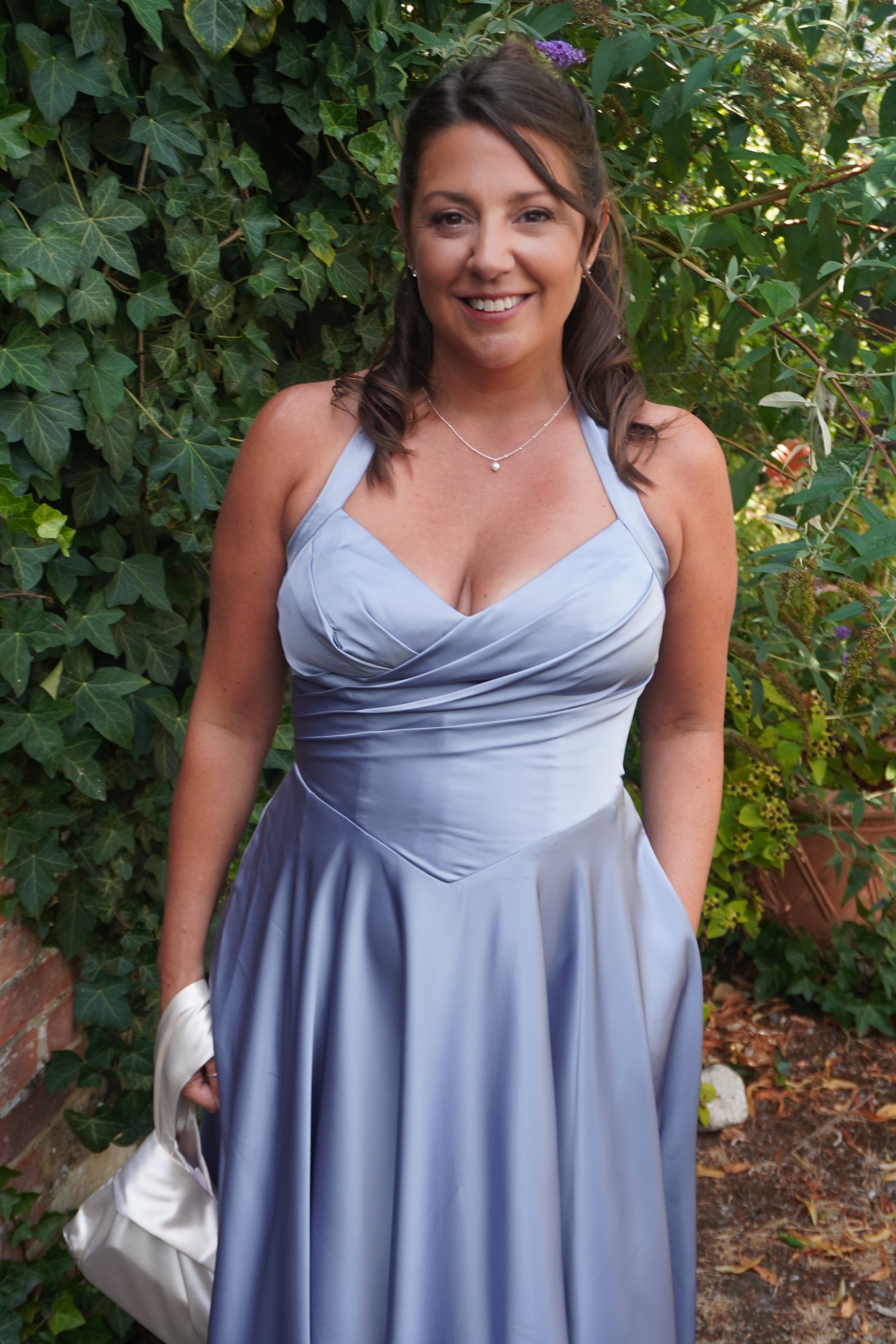 A woman in a light blue dress smiling outdoors in front of green leafy plants.