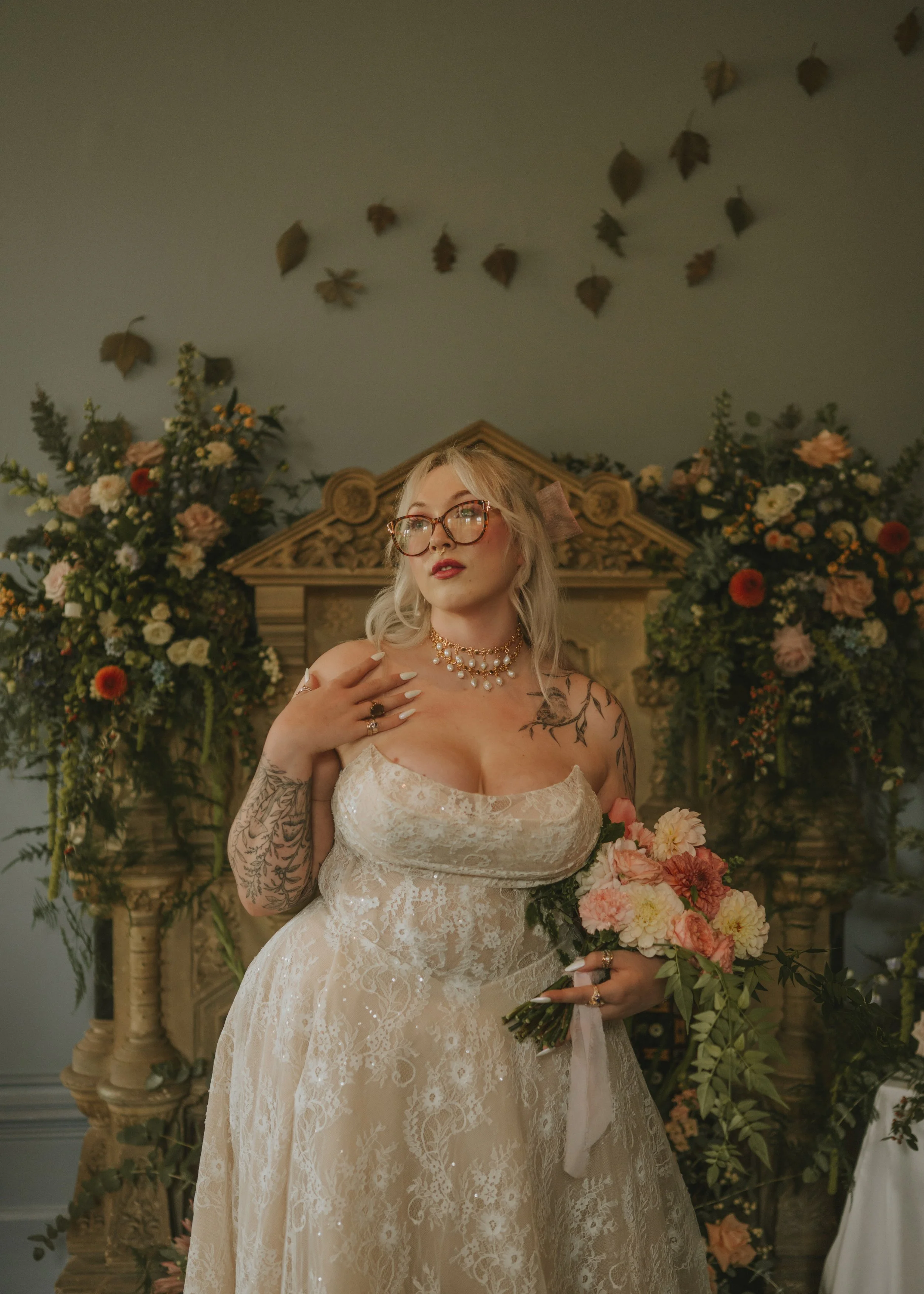 A woman in a lace wedding dress holding a bouquet of flowers, standing in front of a fireplace decorated with flowers and greenery.