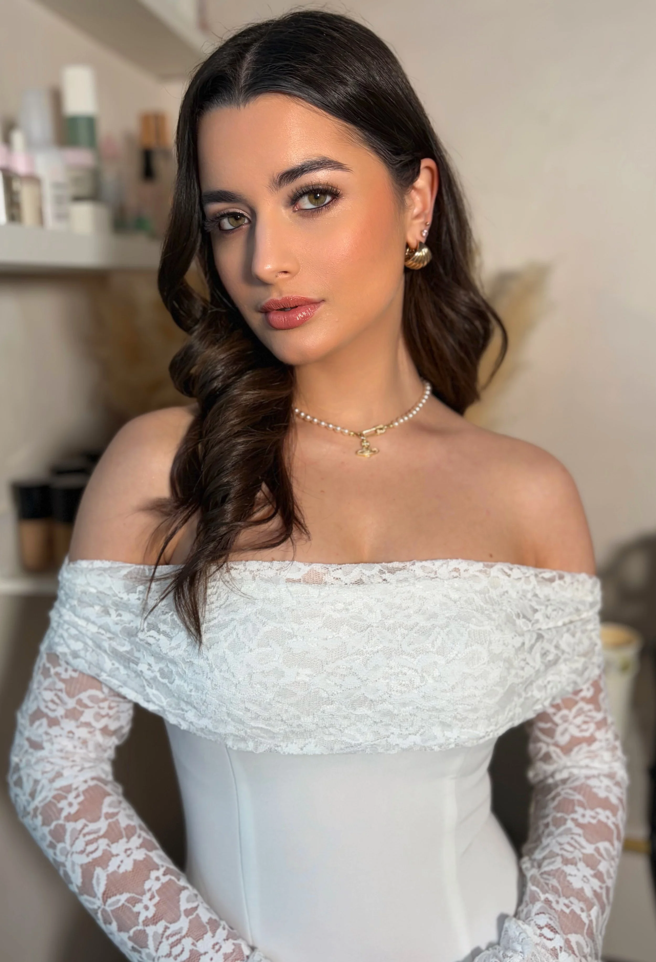 Close-up of a woman with long dark hair, wearing pearl and gold earrings, a pearl necklace with a gold pendant, and an off-shoulder white lace dress, standing indoors with a neutral background.