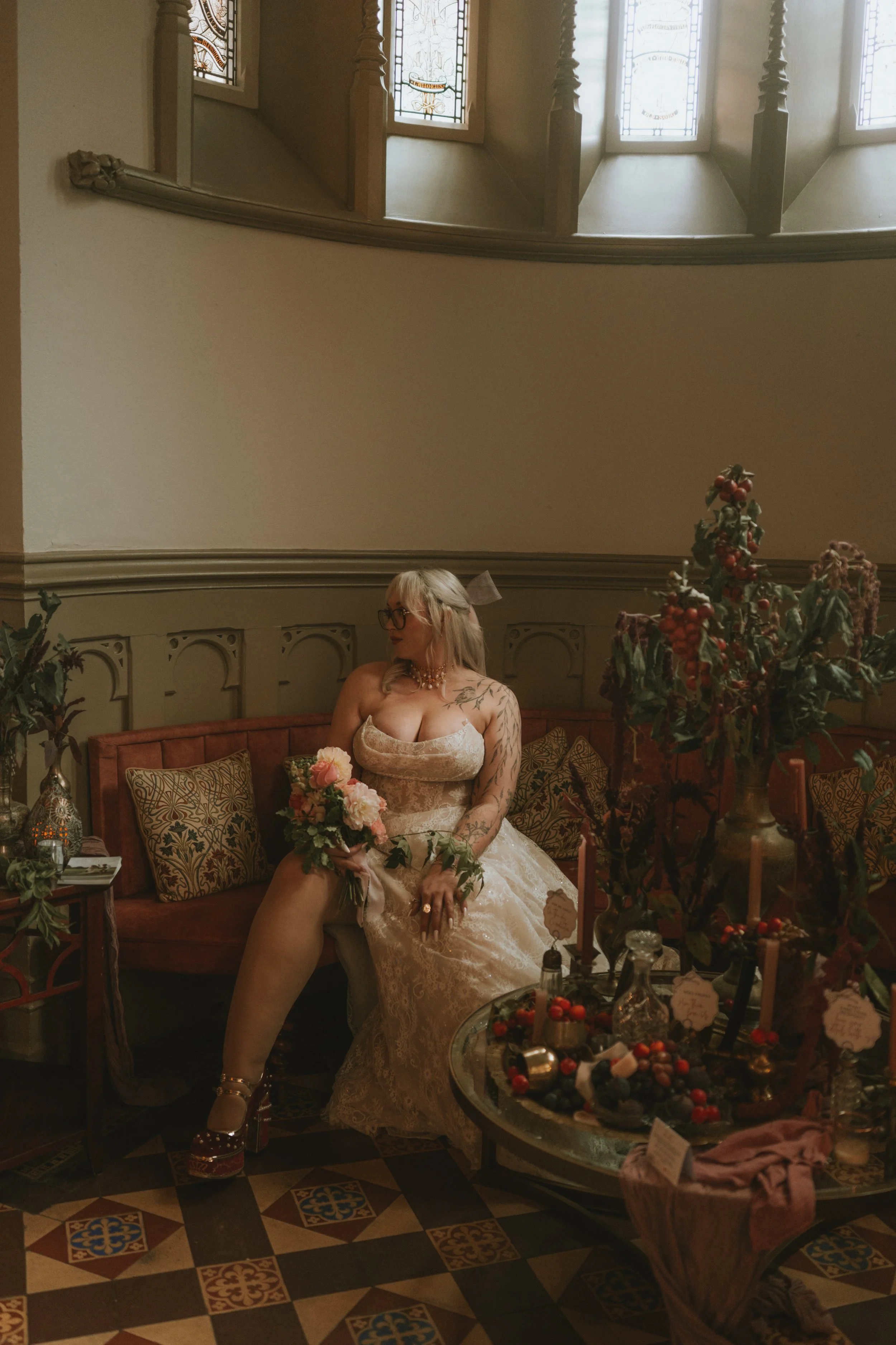 A woman wearing a vintage wedding dress with lace details, glasses, and a bow in her hair, sitting on a vintage sofa with patterned pillows, holding a bouquet of pink and white flowers, in a decorated interior with flowers and candles.