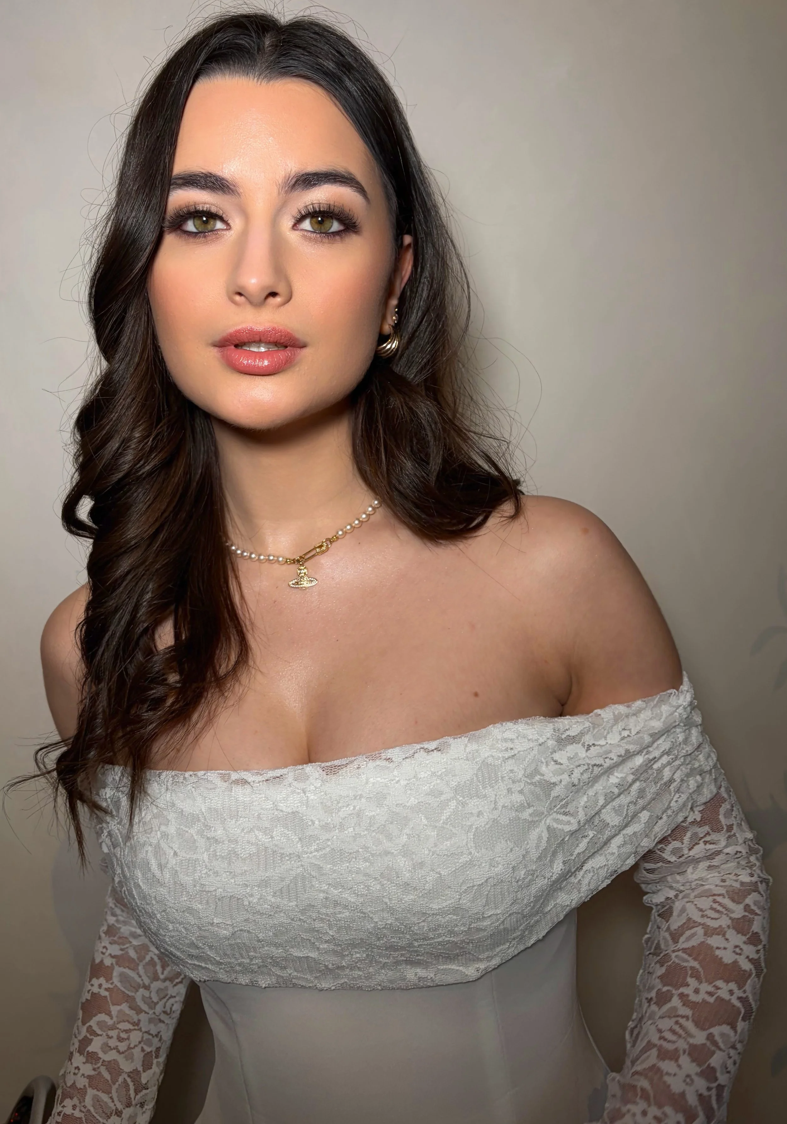 A woman with long dark hair, hazel eyes, and makeup, wearing gold earrings, a pearl necklace with a gold charm, and an off-the-shoulder white lace dress, standing against a plain wall.