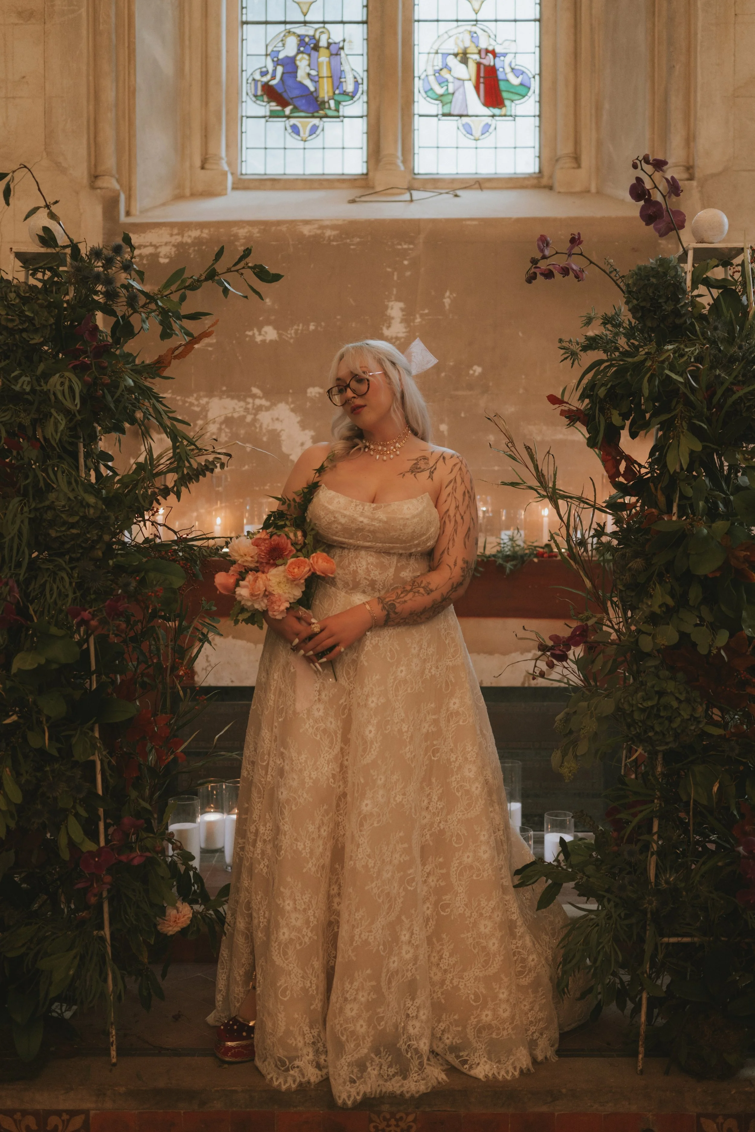 A woman in a lace wedding dress holding a bouquet of flowers standing inside a church with stained glass windows behind her, surrounded by greenery and candles.