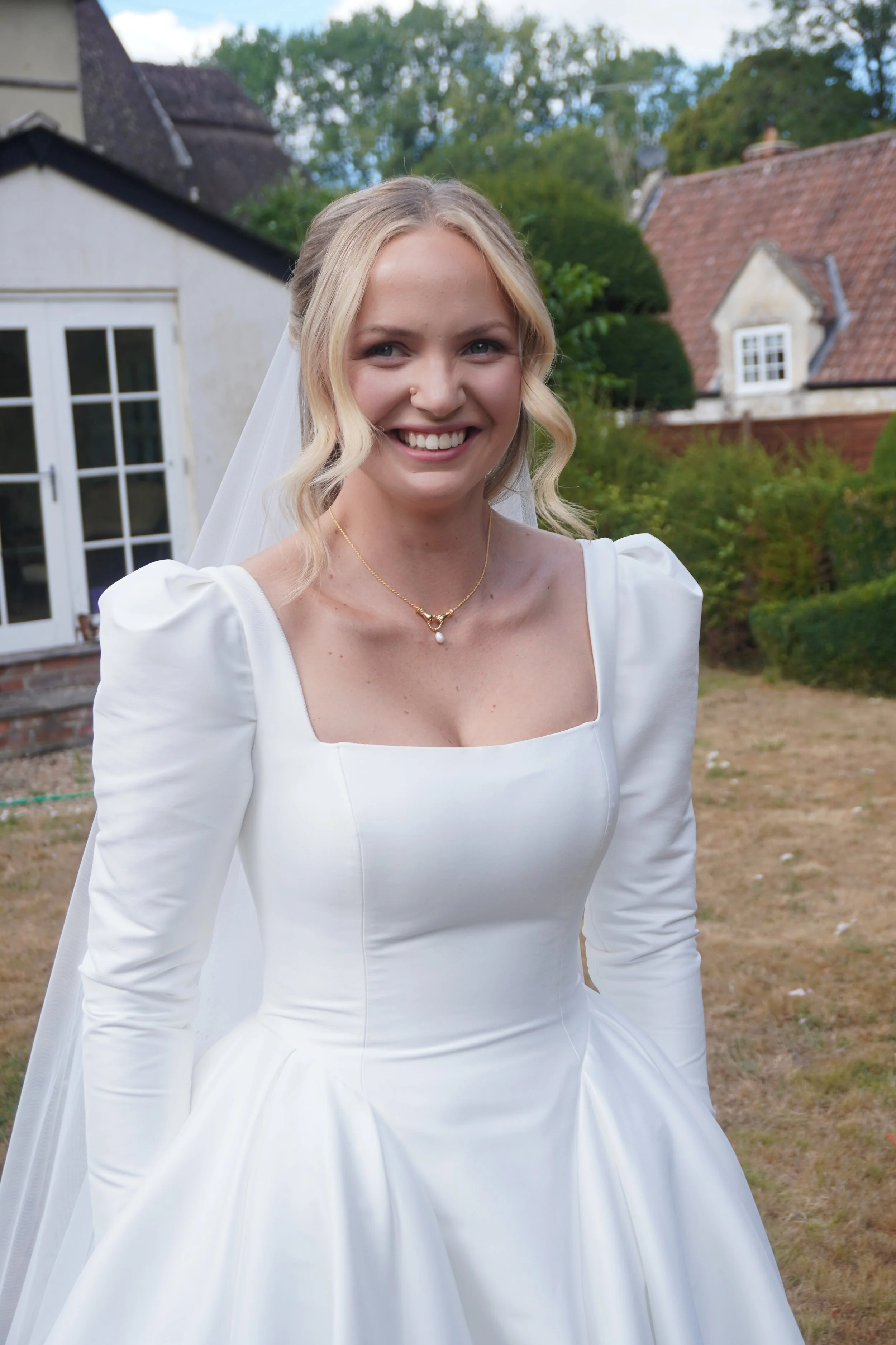 A smiling woman dressed in a white wedding gown with puffed sleeves, standing outdoors in a garden area with houses and trees in the background.