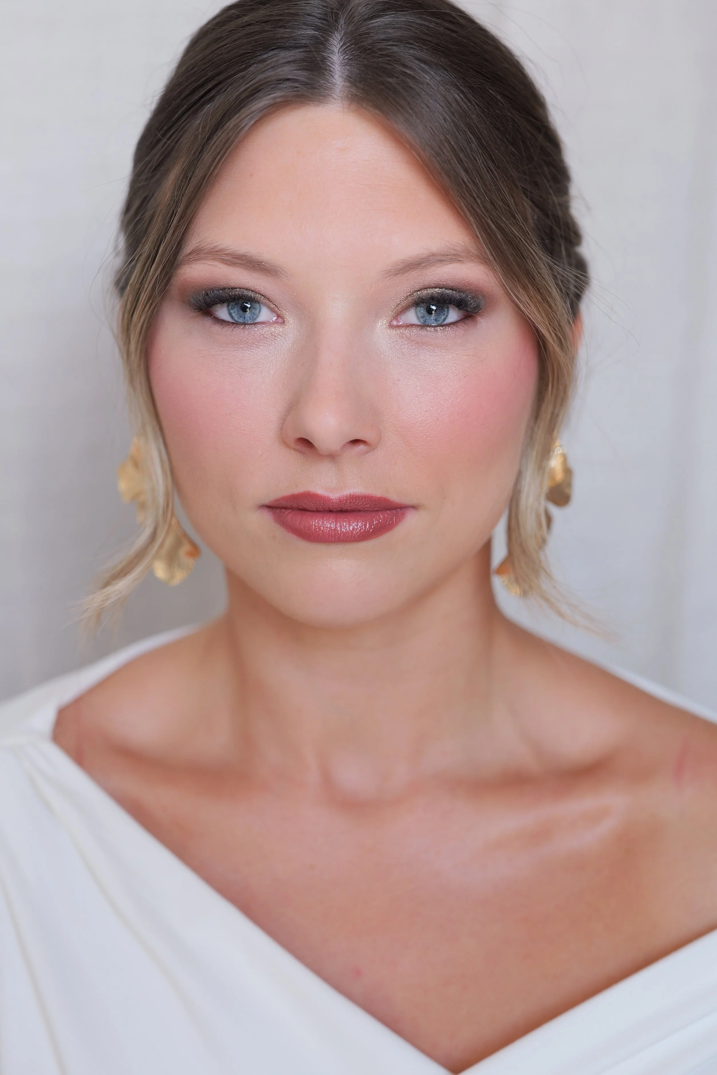 Close-up of a woman with blue eyes, light makeup, and light brown hair styled in loose waves, wearing gold earrings and a white top.