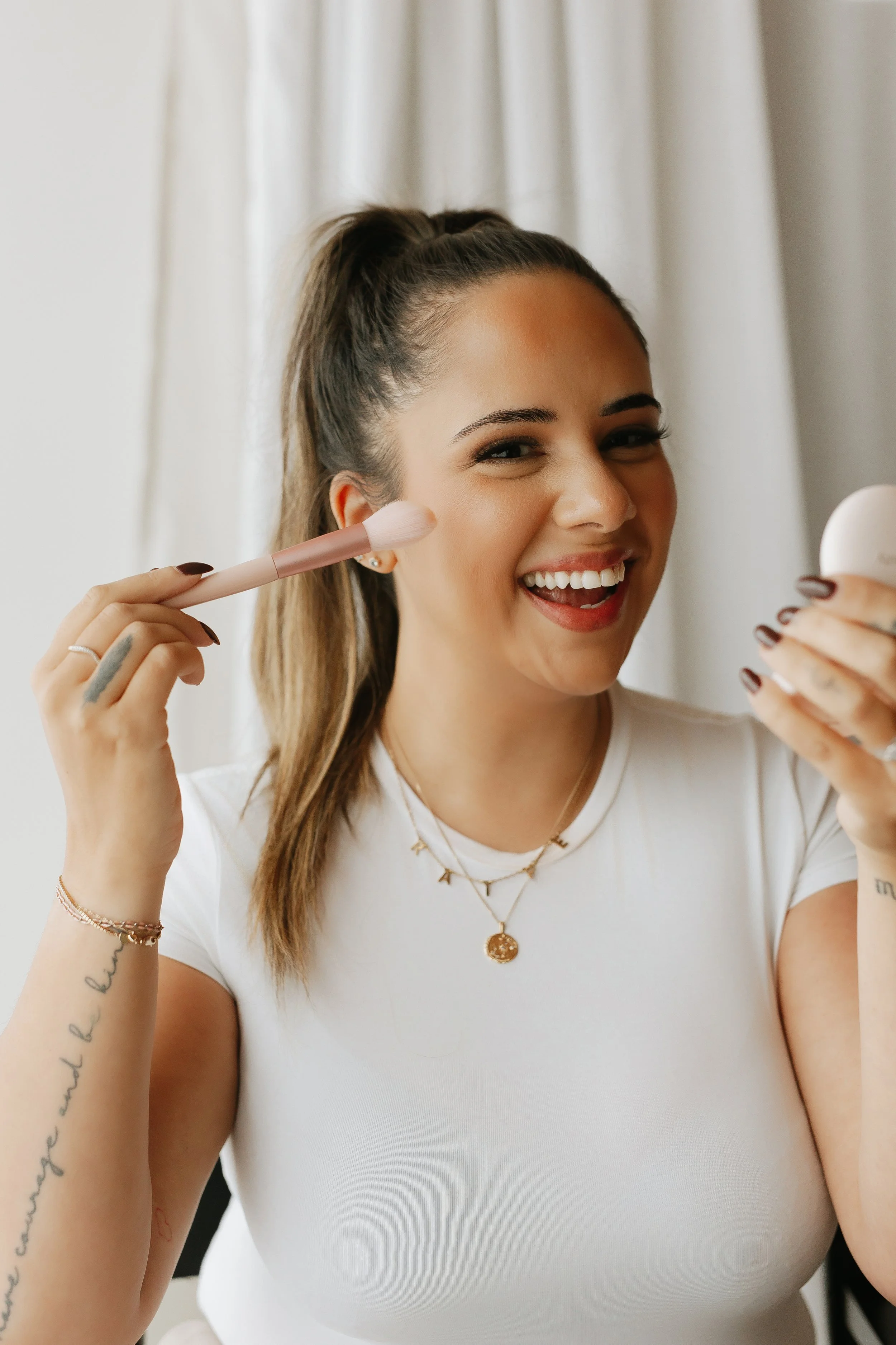 A woman smiling while holding a mirror in one hand and applying makeup with a brush in the other. She has her hair tied back and is wearing a white t-shirt and layered necklaces, standing in front of a light-colored curtain.