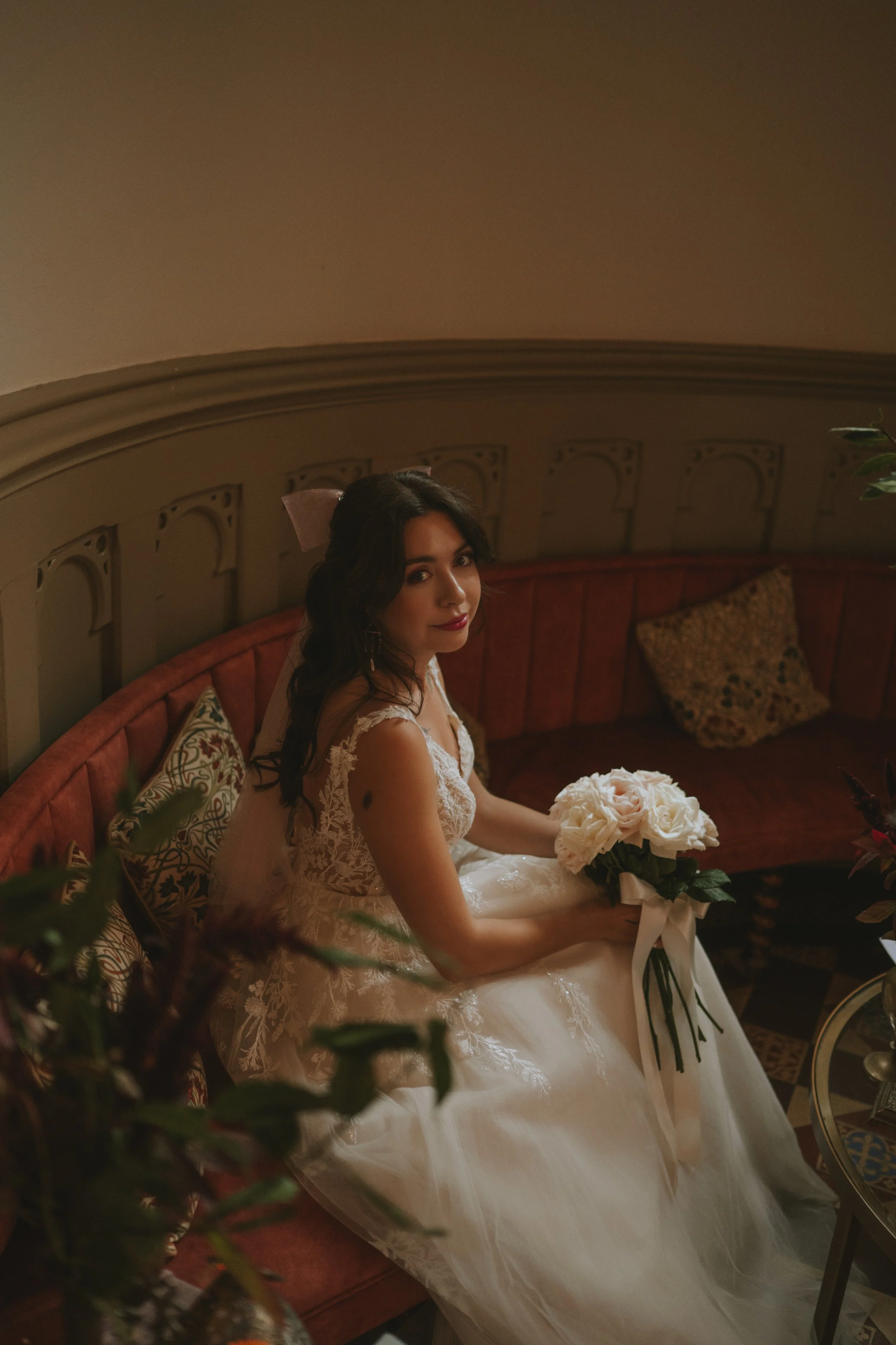 Bride in a wedding dress sitting on a vintage red velvet settee holding a bouquet of white roses, looking at the camera in a warmly lit room.