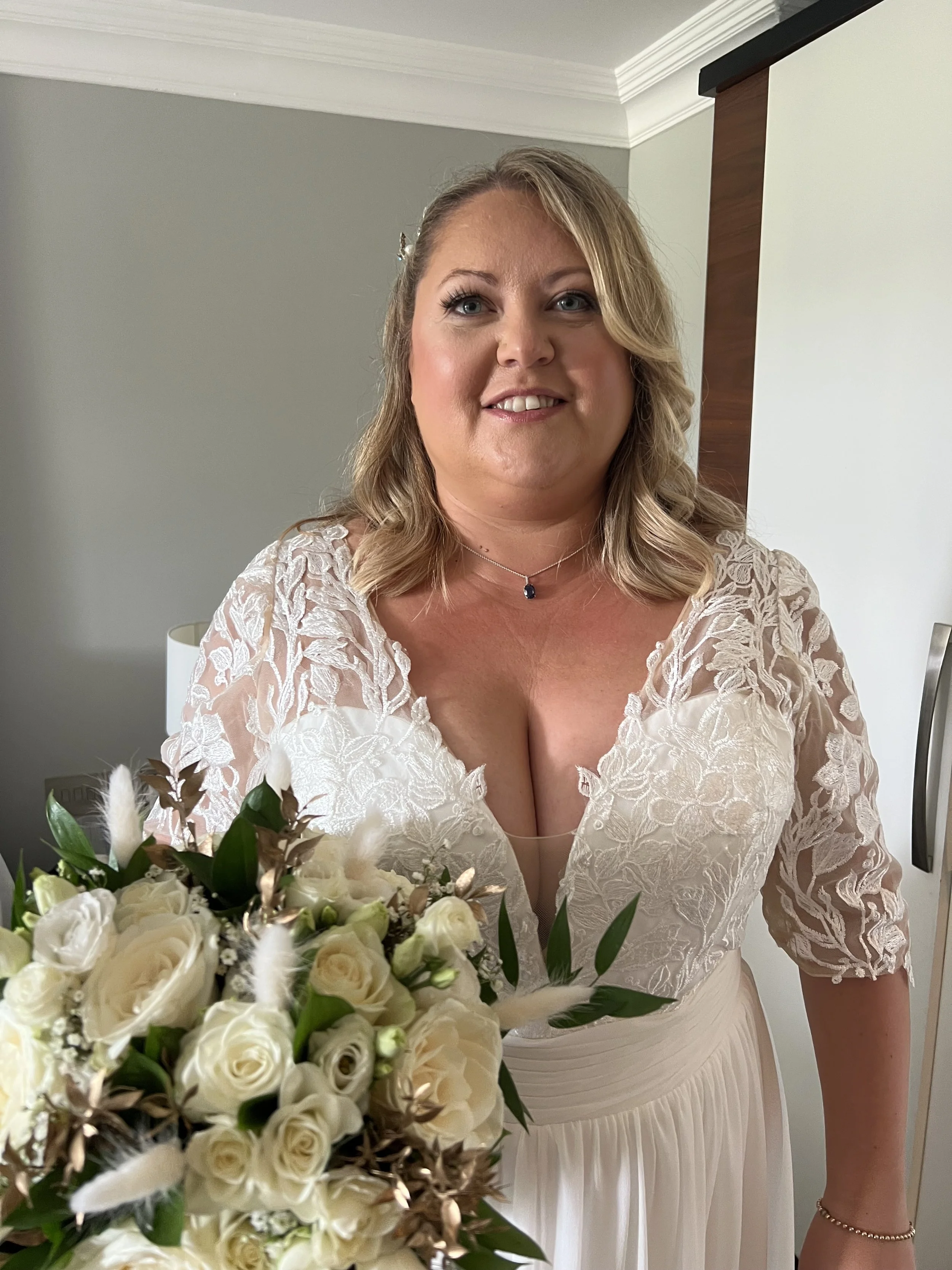 A woman in a wedding dress holding a bouquet of white roses and greenery, smiling indoors.