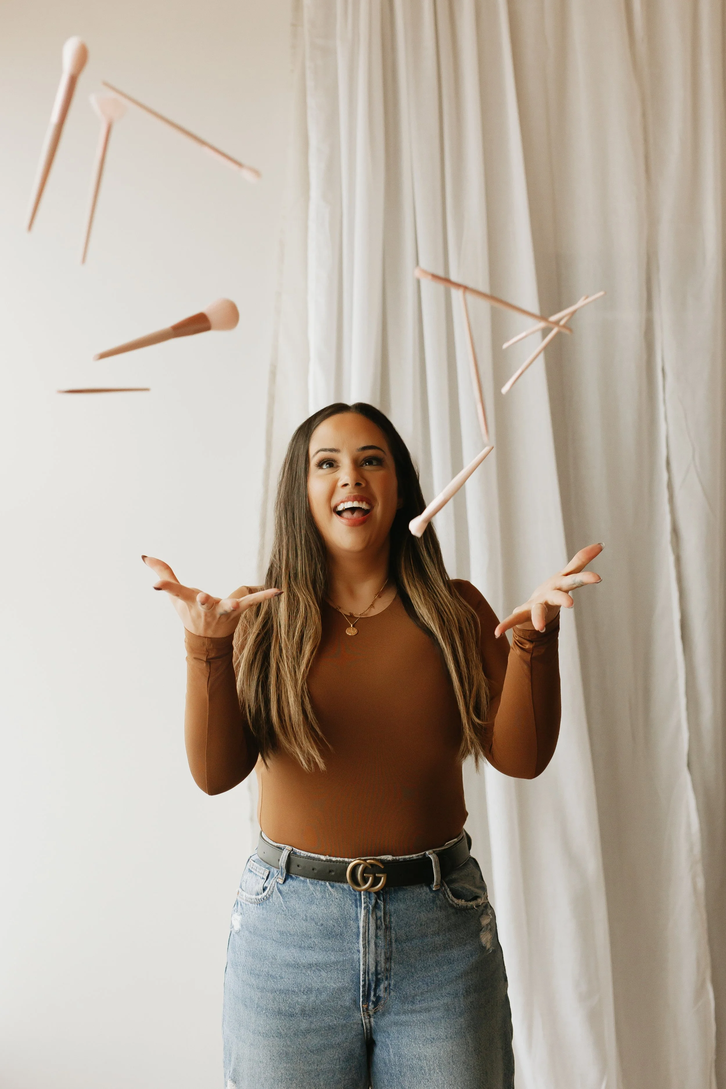 A woman with long brown hair, wearing a brown long-sleeve top and high-waisted jeans, is smiling and throwing makeup brushes into the air indoors near white curtains.