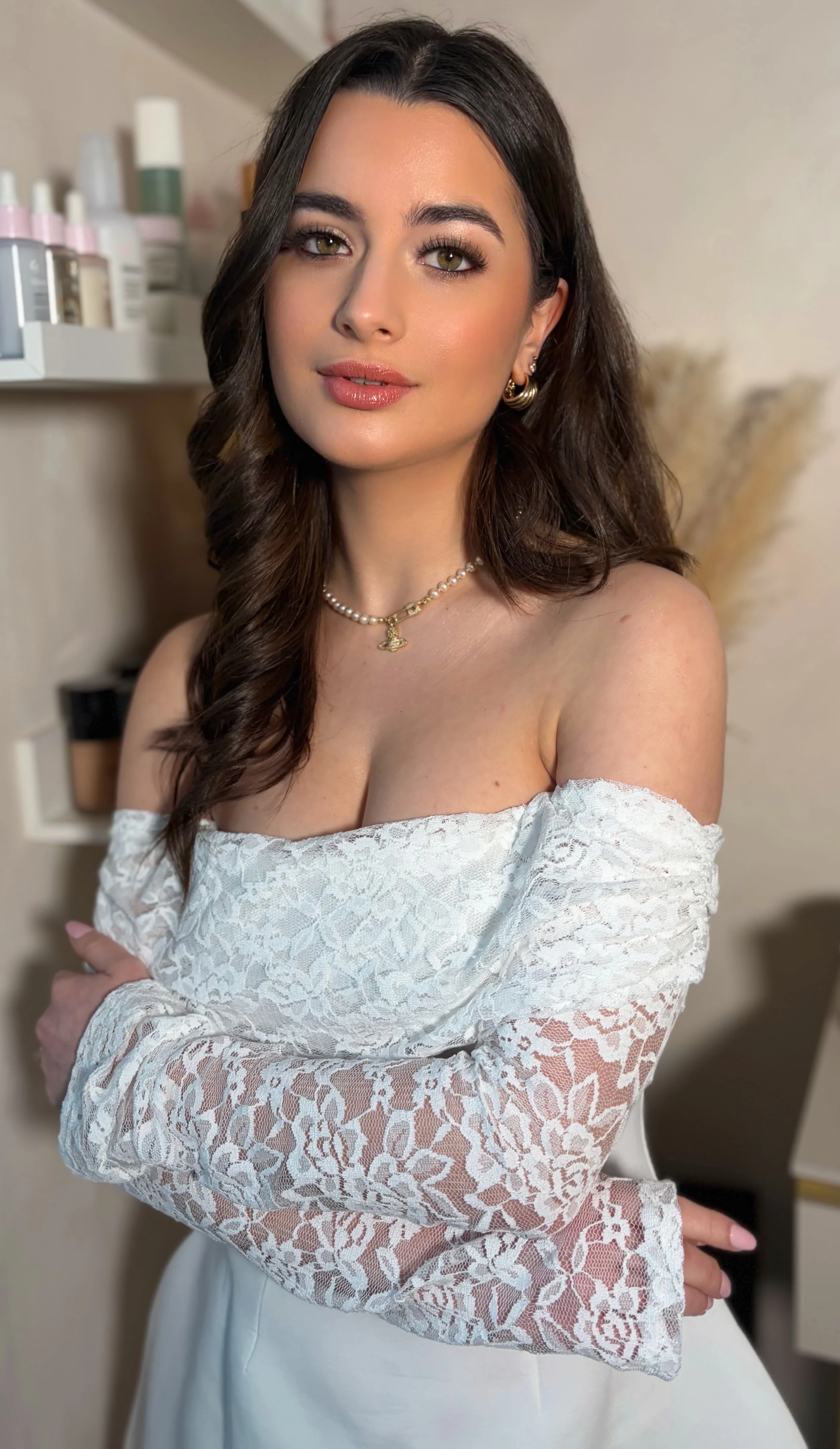 A woman with long, wavy brown hair and light green eyes posing indoors wearing an off-shoulder white lace top, pearl necklace, and gold hoop earrings, with a background of shelves with skincare products.