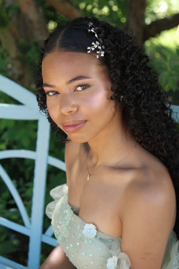 Portrait of a woman with dark curly hair wearing a light-colored strapless dress with floral embellishments, accessorized with a delicate necklace and hair ornament, outdoors with green foliage in the background.