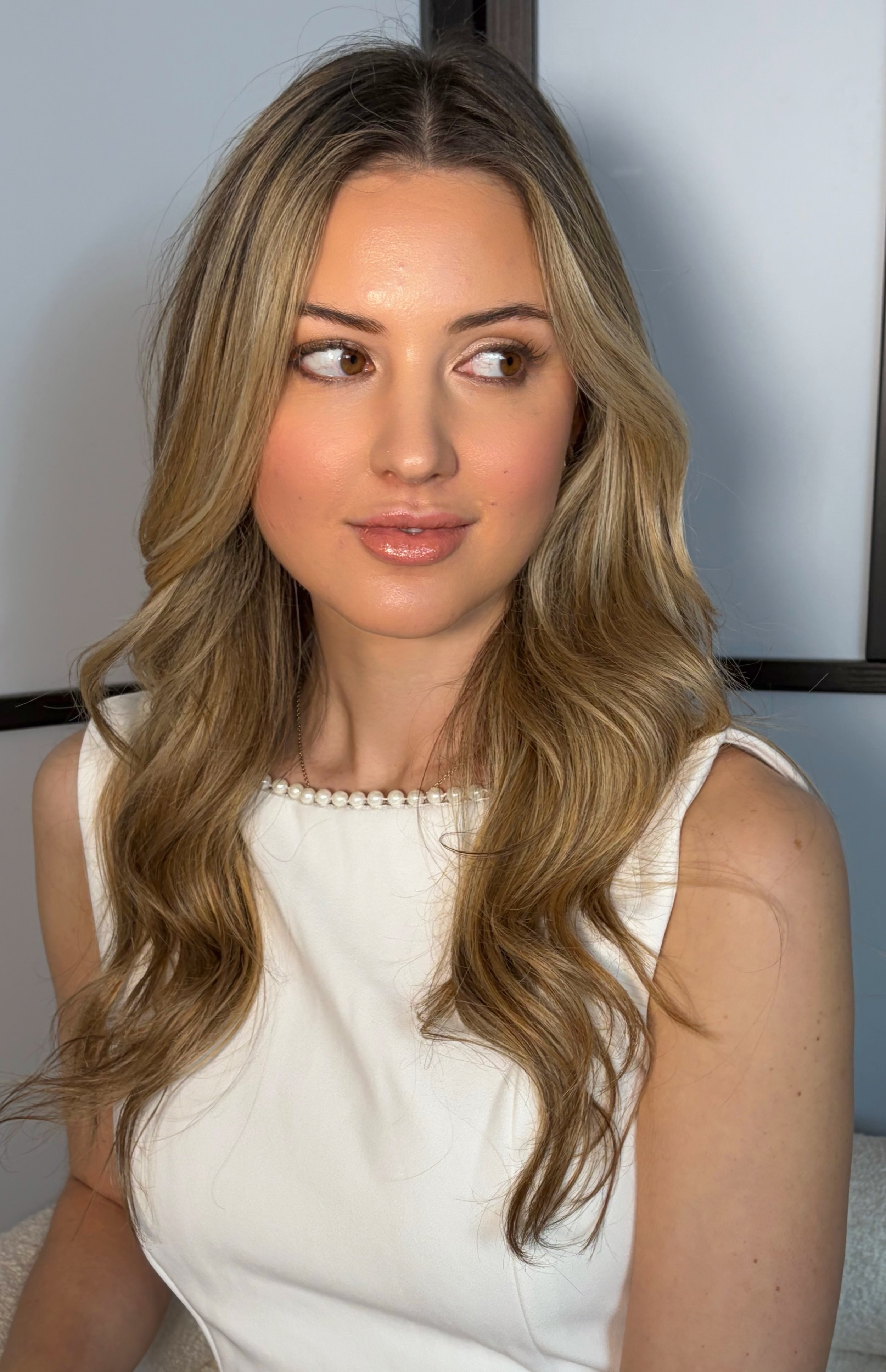 Close-up of a woman with long wavy blonde hair, wearing a white sleeveless dress with a pearl necklace, looking to her right with a neutral expression.